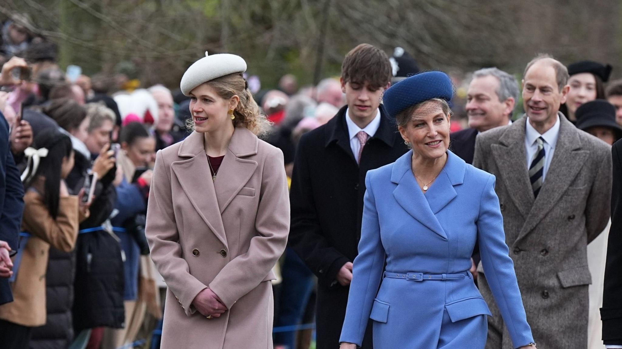 Lady Louise Windsor wearing a grey-pink coat and white hat walks next to the Duchess of York wearing a powder blue coat and dark blue hat. The Earl of Wessex wearing a black coat and pink tie and the Duke of Edinburgh wearing a brown coat and striped tie walk behind them.