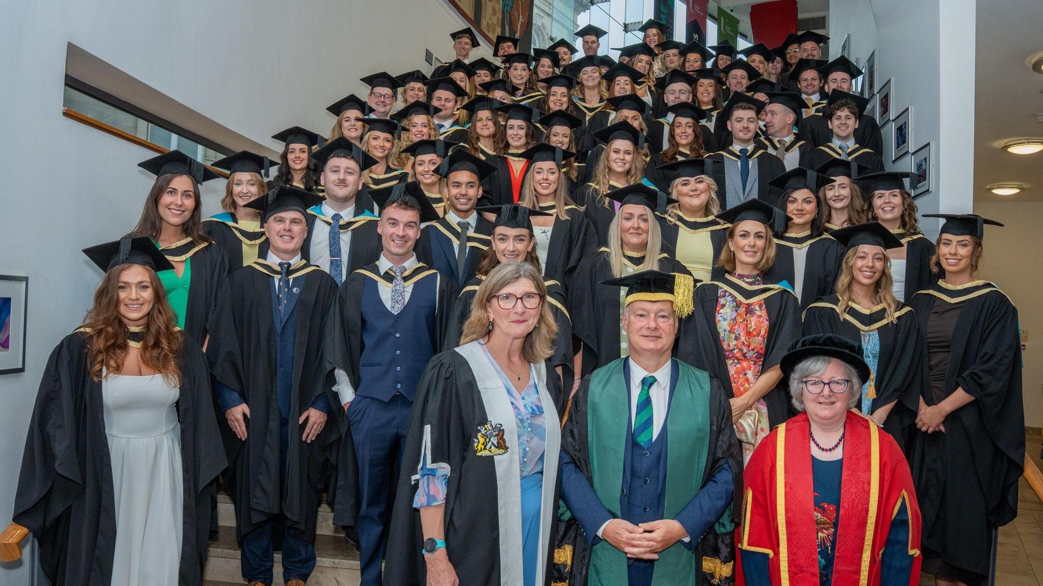 a group of 69 medical graduates stand on the stairs of Derry's millenium forum following their gradution. the men and women are wearing formal dress and mortar boards and gons