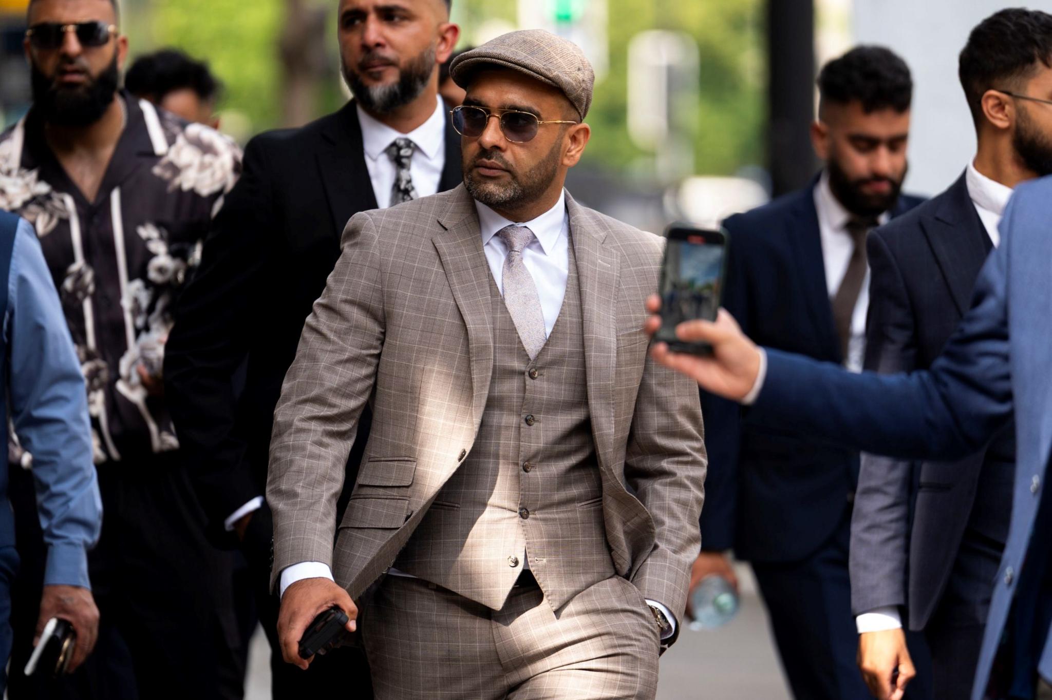 A man in a beige three-piece suit and a beige cap walks outside, surrounded by other men in suits. The man is wearing sunglasses and he has a neatly trimmed black beard.