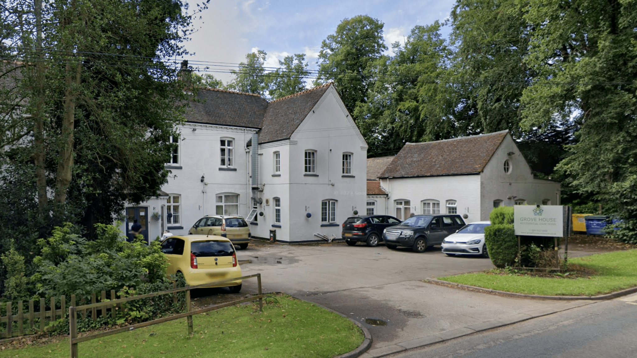 The outside of Grove House care home with cars parked on the drive and green trees surrounding the building.