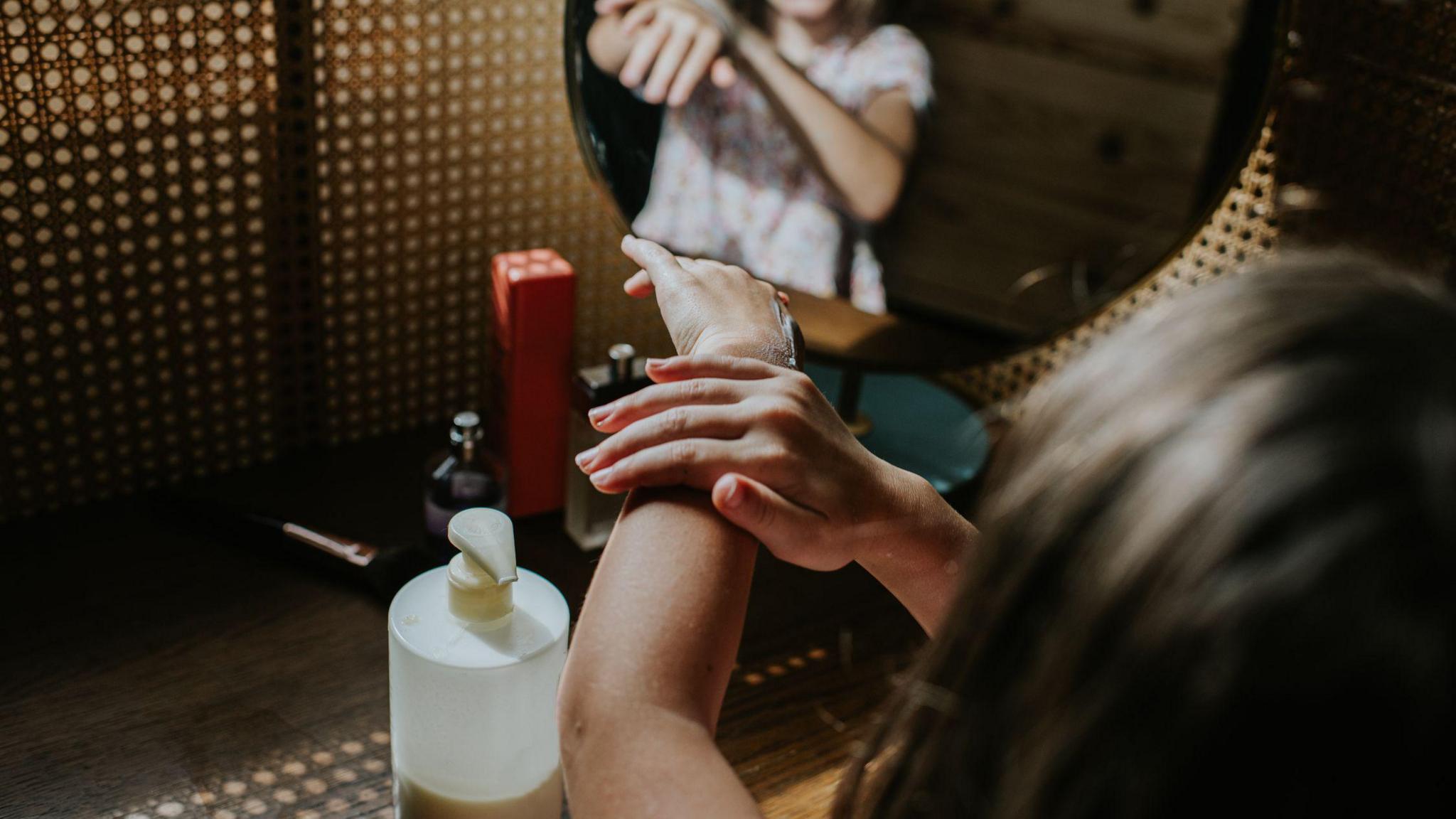 Stock image of a teenager rubbing cream into her hands, with a half-empty bottle on a table in front of her, and a mirror in the background.