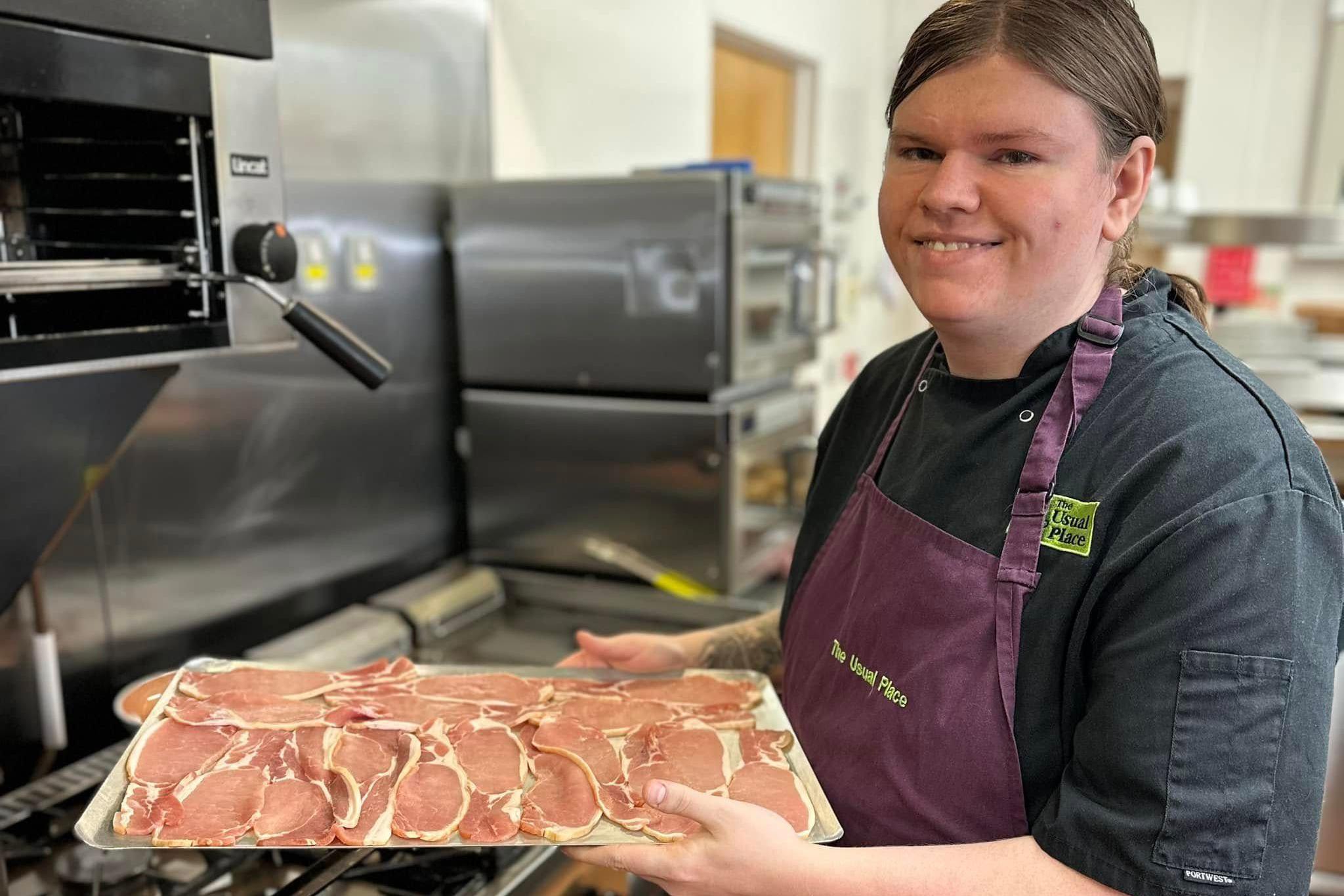 A young woman with dark hair in a black T-Shirt and purple apron holds a large tray of bacon which is about to go under the grill in a large commercial kitchen