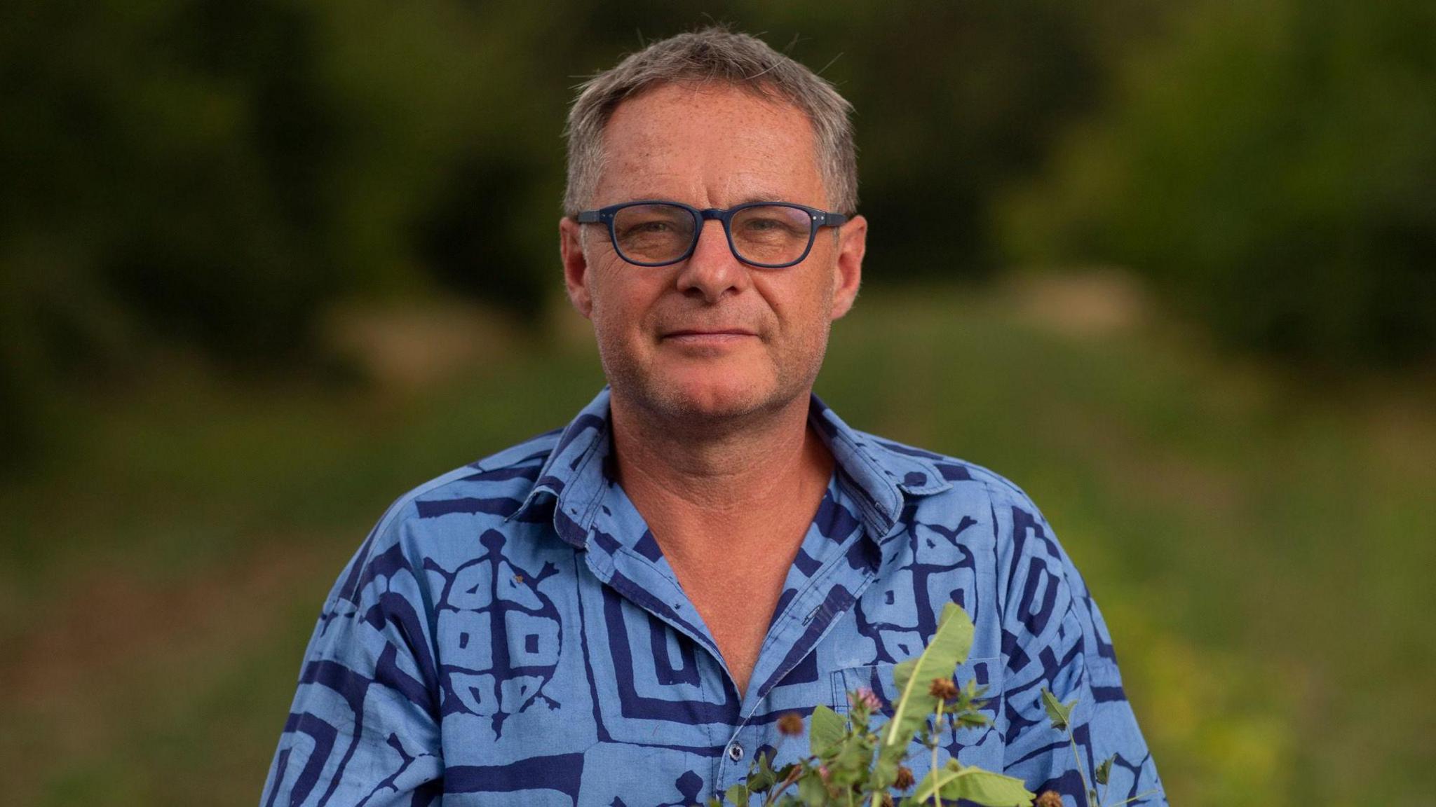 David Wolfe is pictured smiling to camera while standing in a field. He has short grey hair and is wearing a blue shirt with patterns on it. He is wearing dark coloured glasses and he is holding a plant.