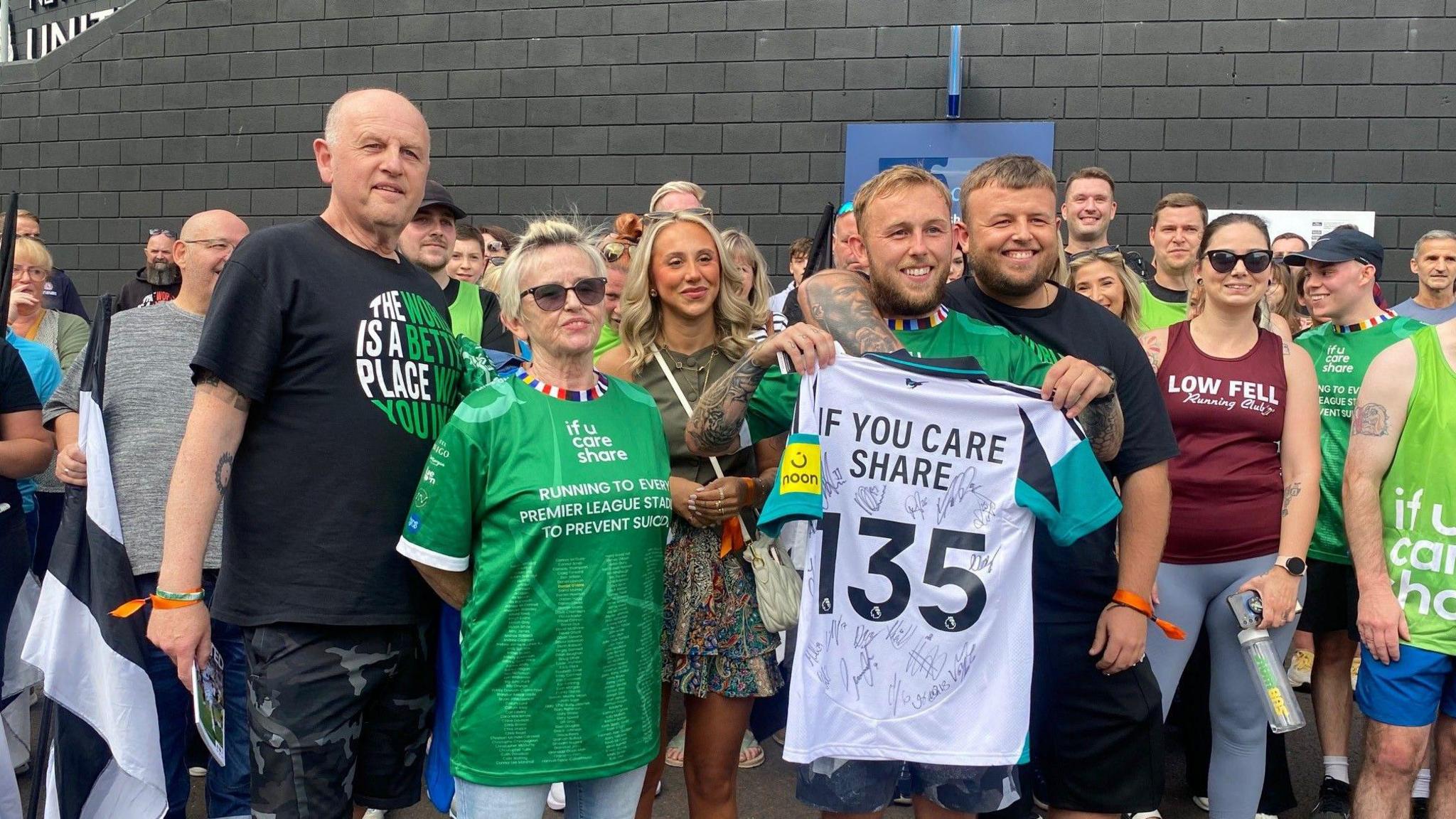 Matthew Smith holding a signed Newcastle United Shirt. It reads "If You Care Share 135". He stands next to his friends and family and a crowd of supporters.