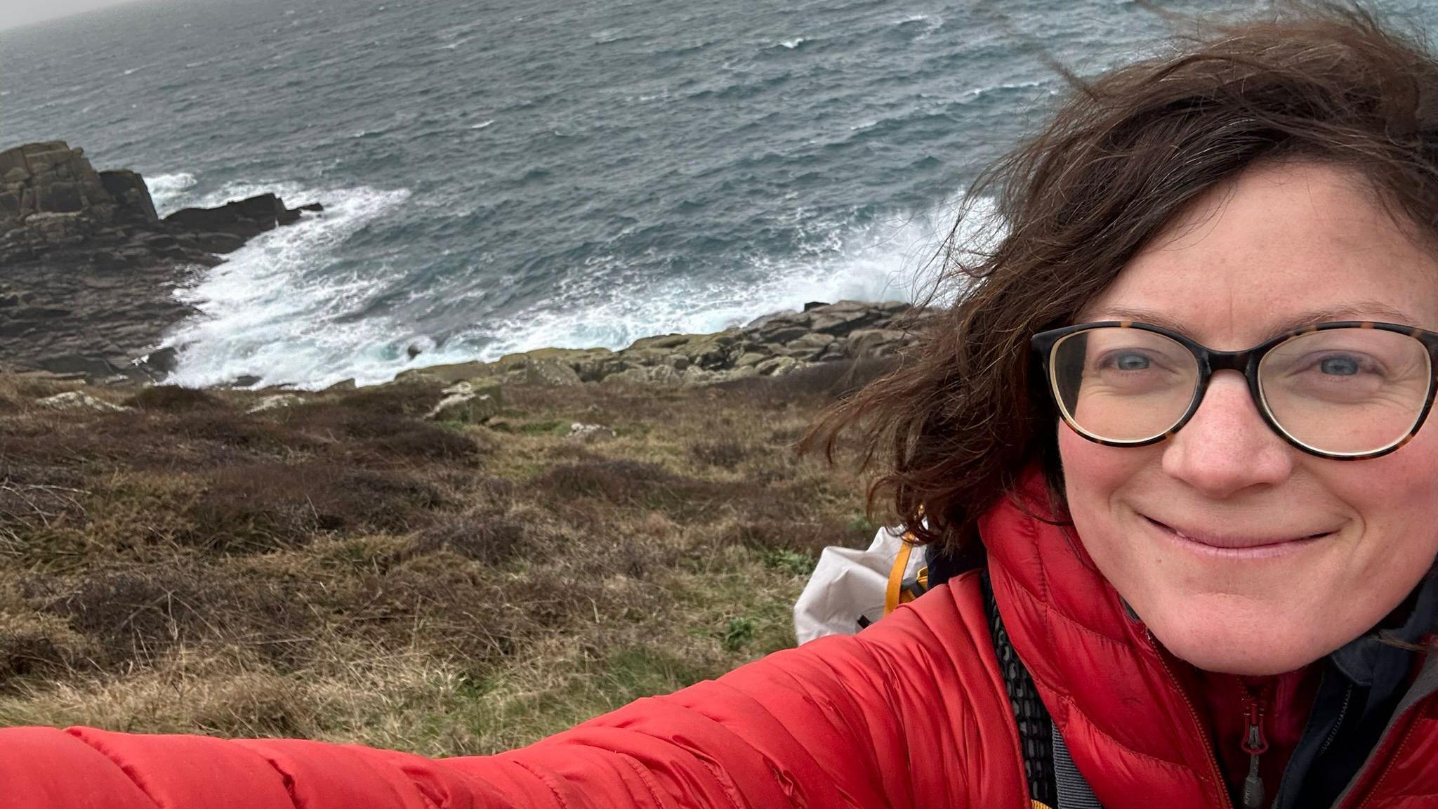 A selfie of Claire Allen wearing a red coat and glasses. Behind her is the sea, bracken and heather. It is a windy day as her hair is blowing and the sea looks rough with lots of sea foam and spray