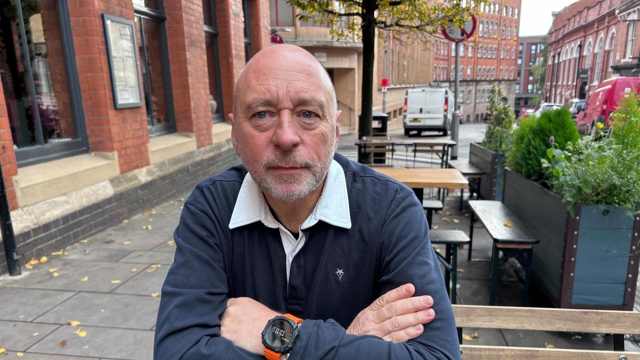 Billy Millward sits on a bench outside a cafe in Northampton town centre. Behind him there are rows of tall red brick buildings. There is a green and modern plant pot to his left, with green plants and trees poking out of it. There are other empty seats and benches behind him. He is wearing a navy and white rugby-style top and a black watch with an orange strap. He has greying stubble on his beard. There is a white van in the distance behind him.
