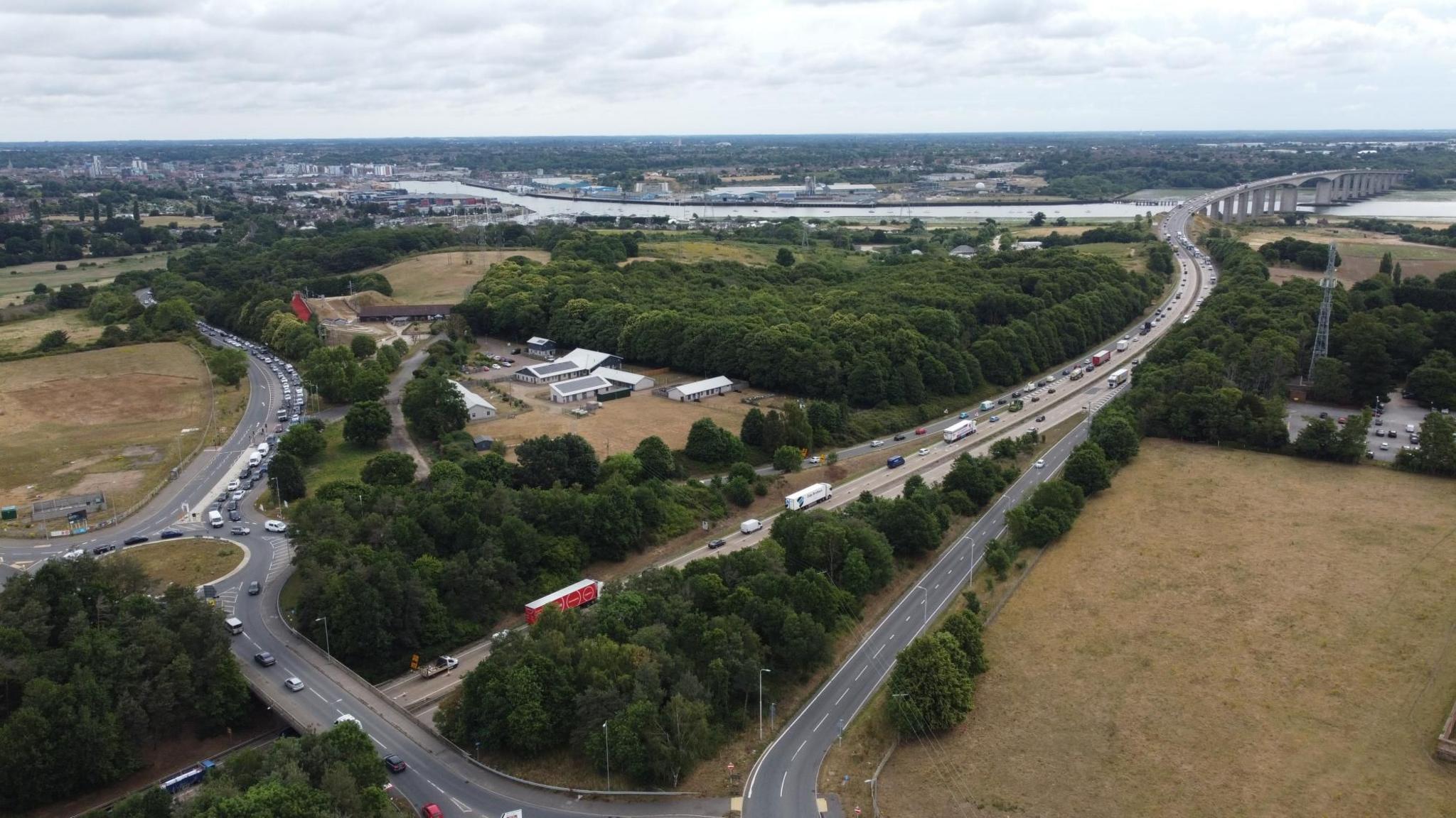 Aerial image of the A14 and Orwell Bridge, taken from above the village of Wherstead. Traffic can be seen queuing/moving slowly as the dual carriageway is cut to one lane each way. Ipswich docks/waterfront is visible upstream, with the town centre in the background.