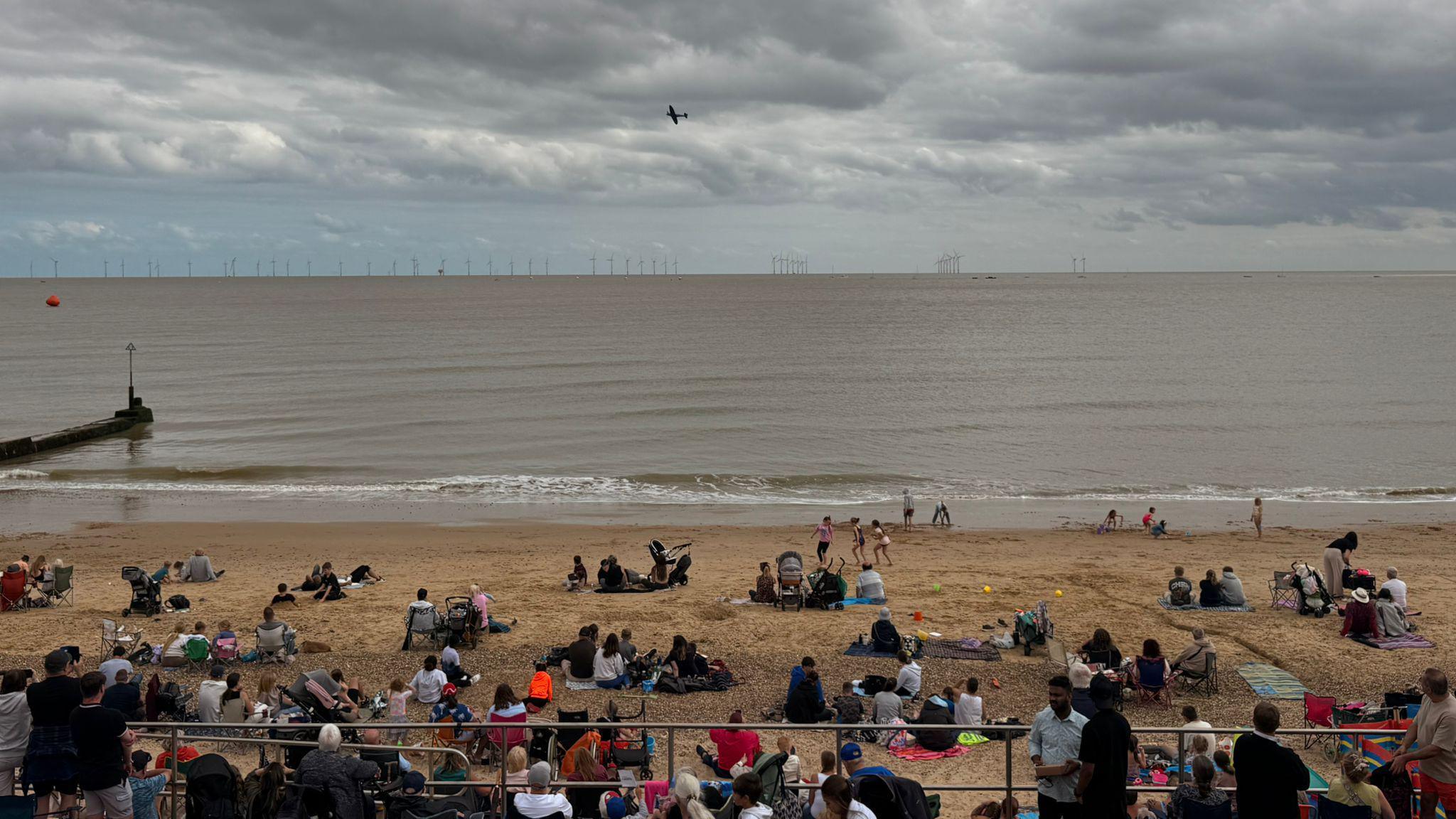 Groups of people sitting on the sandy beach as a plane flies over. In front of them is the North Sea.