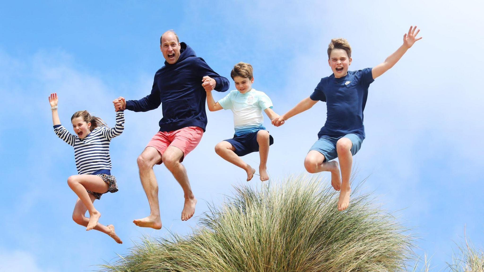 Photograph shows Charlotte with a big smile jumping above a bush while holding the prince of wales' hand, the prince of wales is holding the hand of prince louis and louis is holding prince george's hand