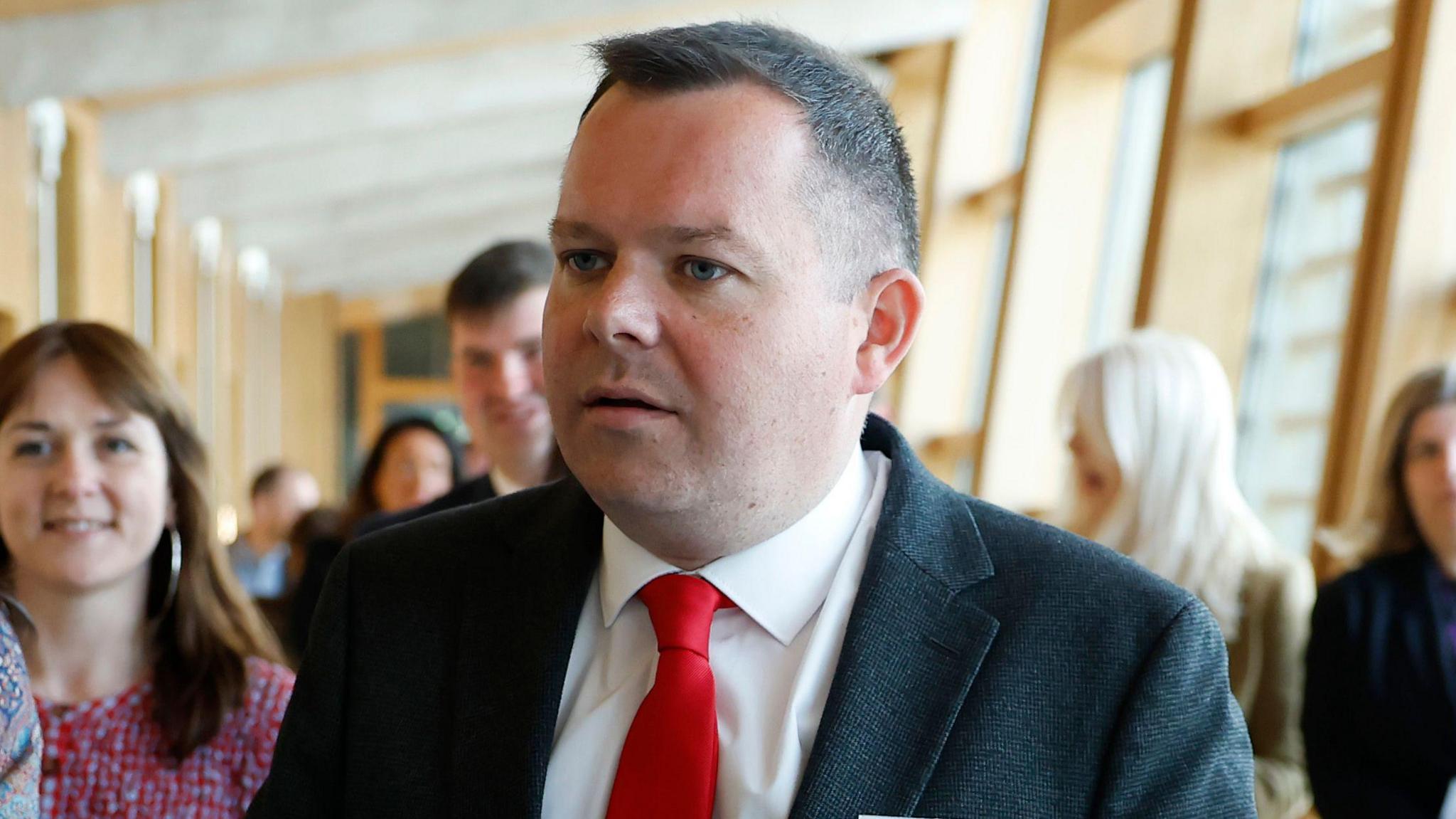 A man with short dark hair, wearing a dark blazer, a white shirt and a red tie, walking into the chamber at Holyrood