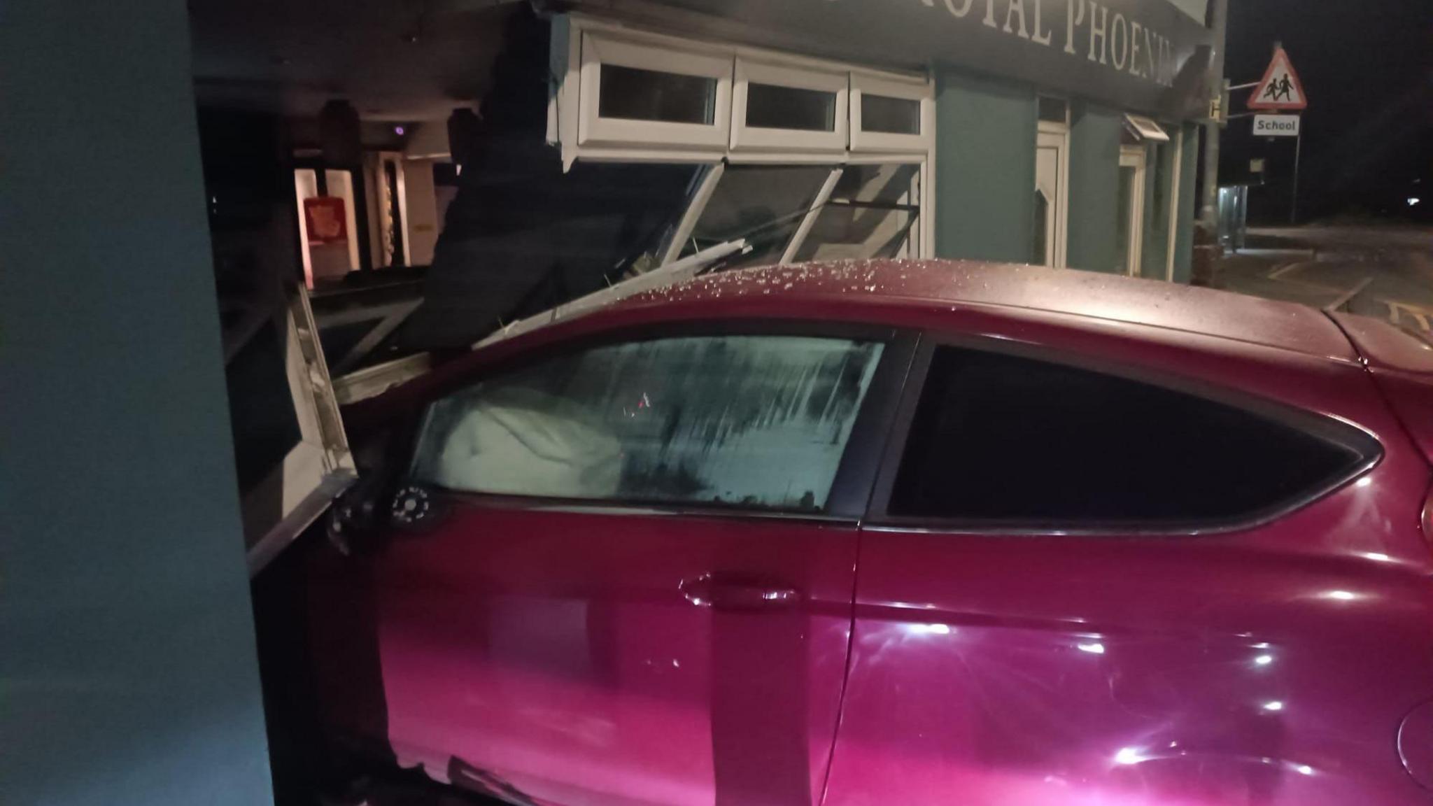 A red car with its front end inside the restaurant - the windows are completely smashed and the car appears to have run over metal barriers that were on the street outside