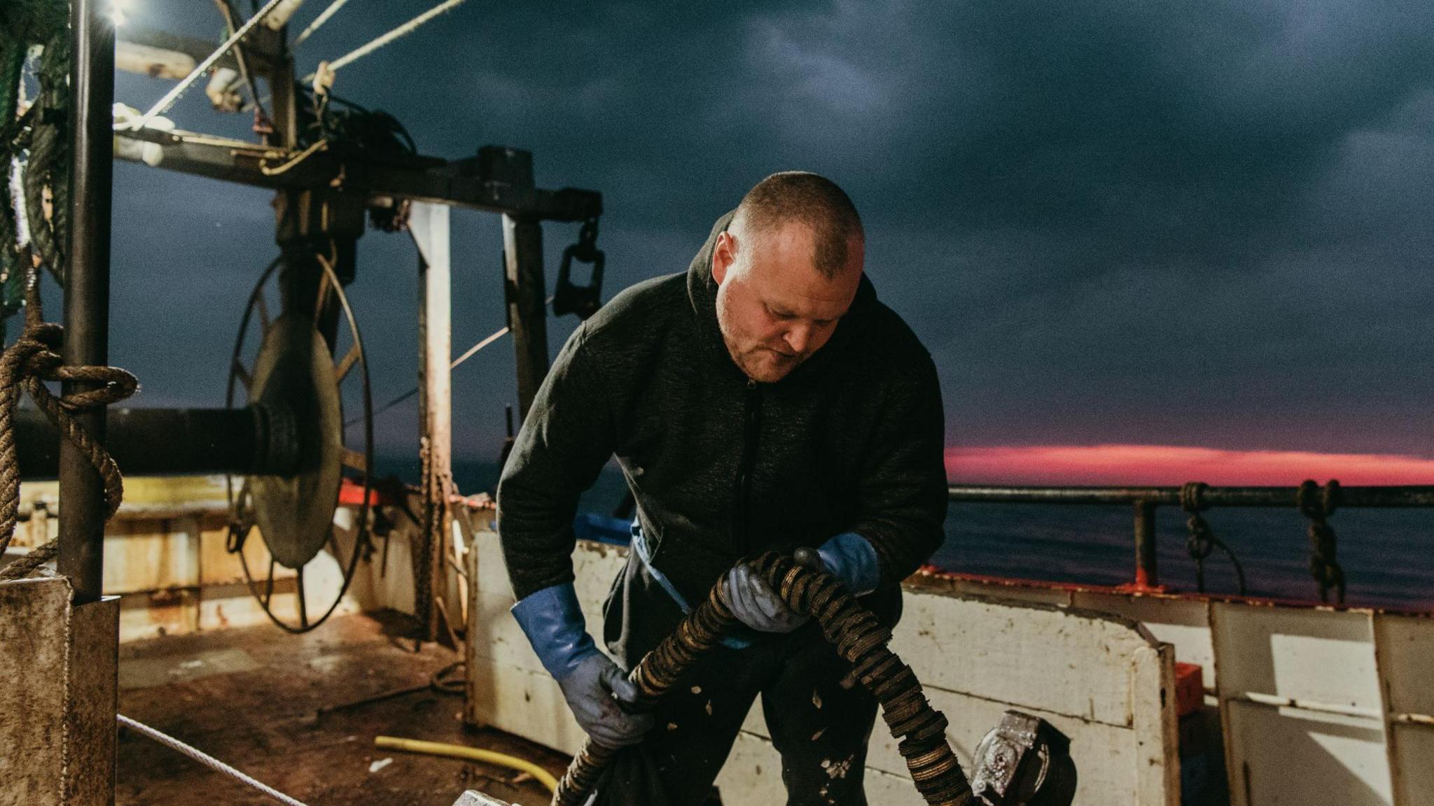 A colour image of a man working on a fishing boat. The sun is rising over his shoulder and the sky on the horizon is a dusky pink. The rest of the sky is dark and filled with clouds. 