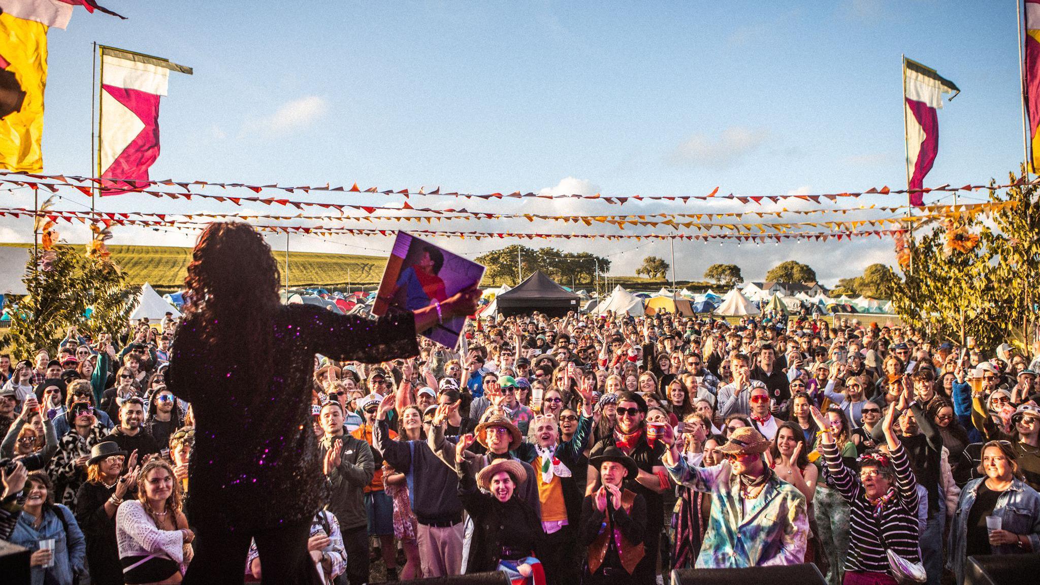 A crowd at the Westival, the photograph taken from behind a performer on stage and showing hundreds of people smiling and clapping, on a sunny day