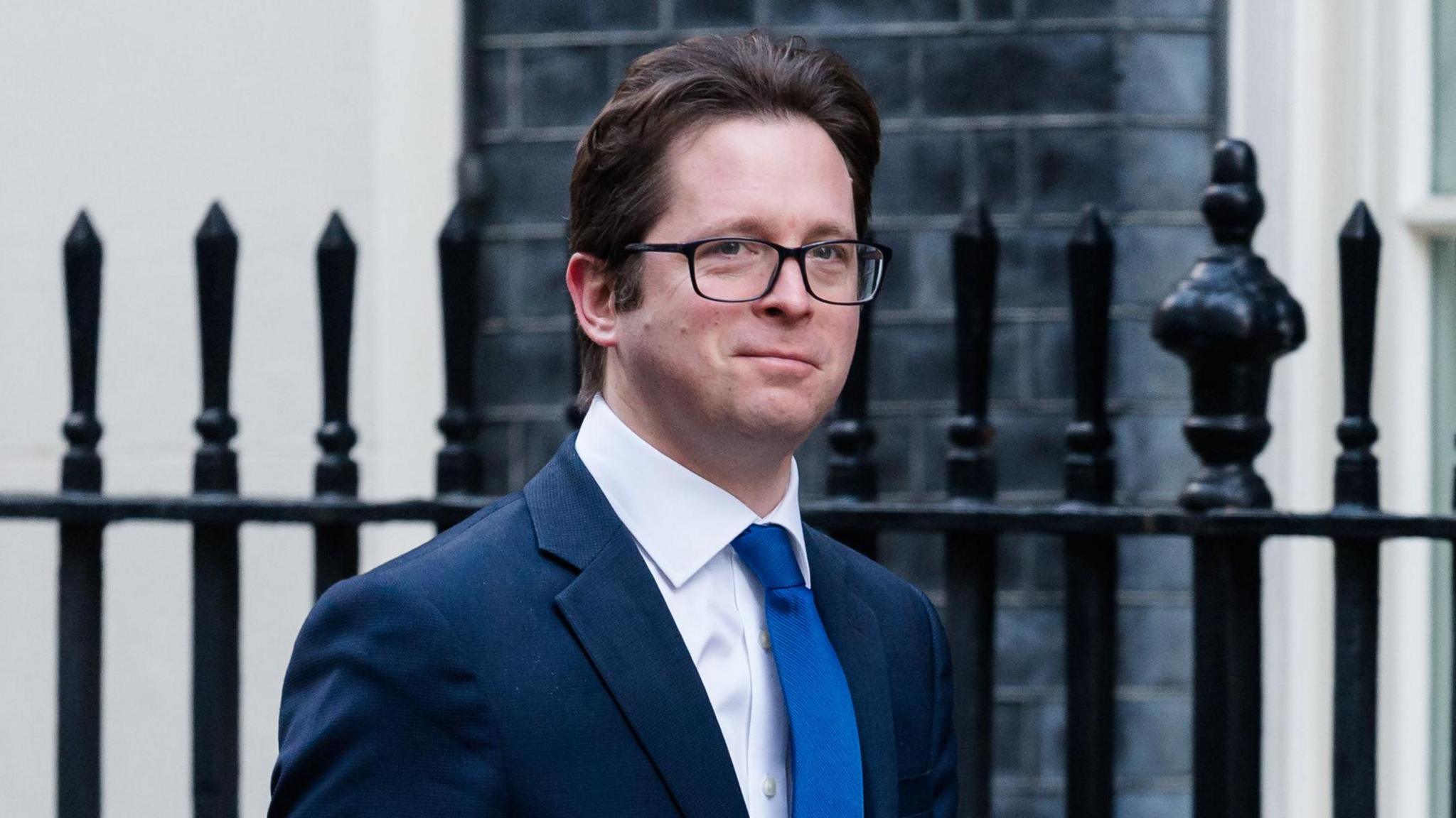 Alex Burghart - a man with brown hair, black square framed glasses wearing a navy suit jacket, a white collared shit and a blue tie. He is walking in front of a black iron fence and a black brick building.