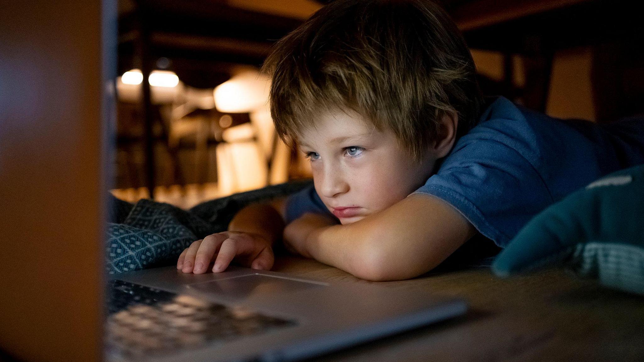 Young boy with blonde coloured hair lying down and looking at a laptop screen 