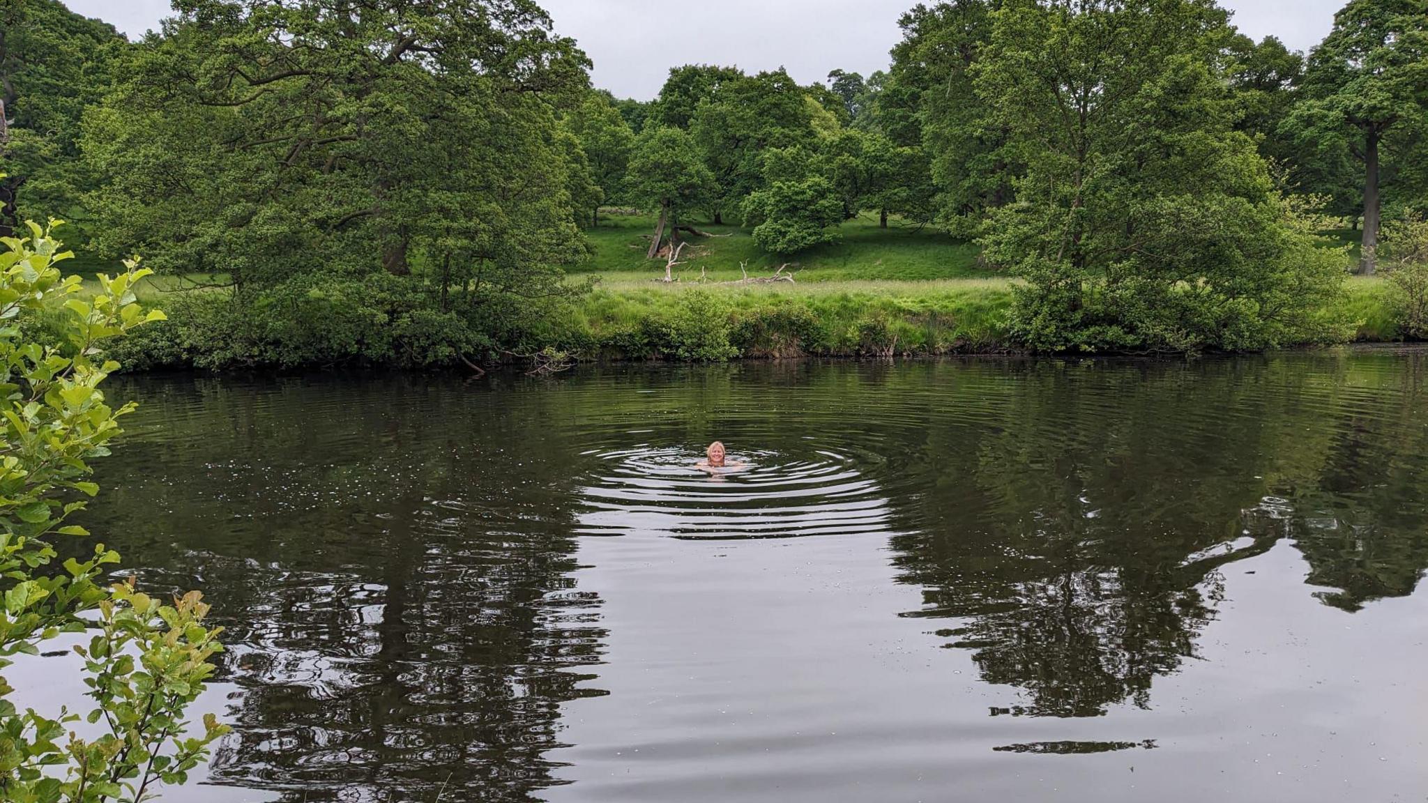 A woman swimming in a river by a tow path with greenery surrounding the bank