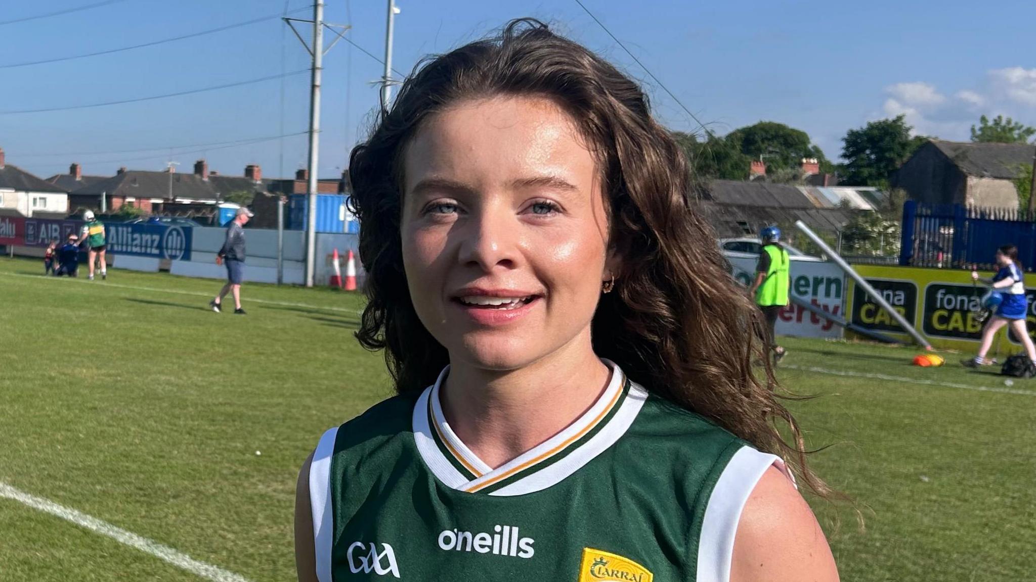 Paula McGivern has long wavy brown hair and is standing on a grass pitch. She is wearing a dark green short sleeved jersey with white and yellow trim. The top says o'neills in white and GAA on one side. A yellow club crest is on the other side.
