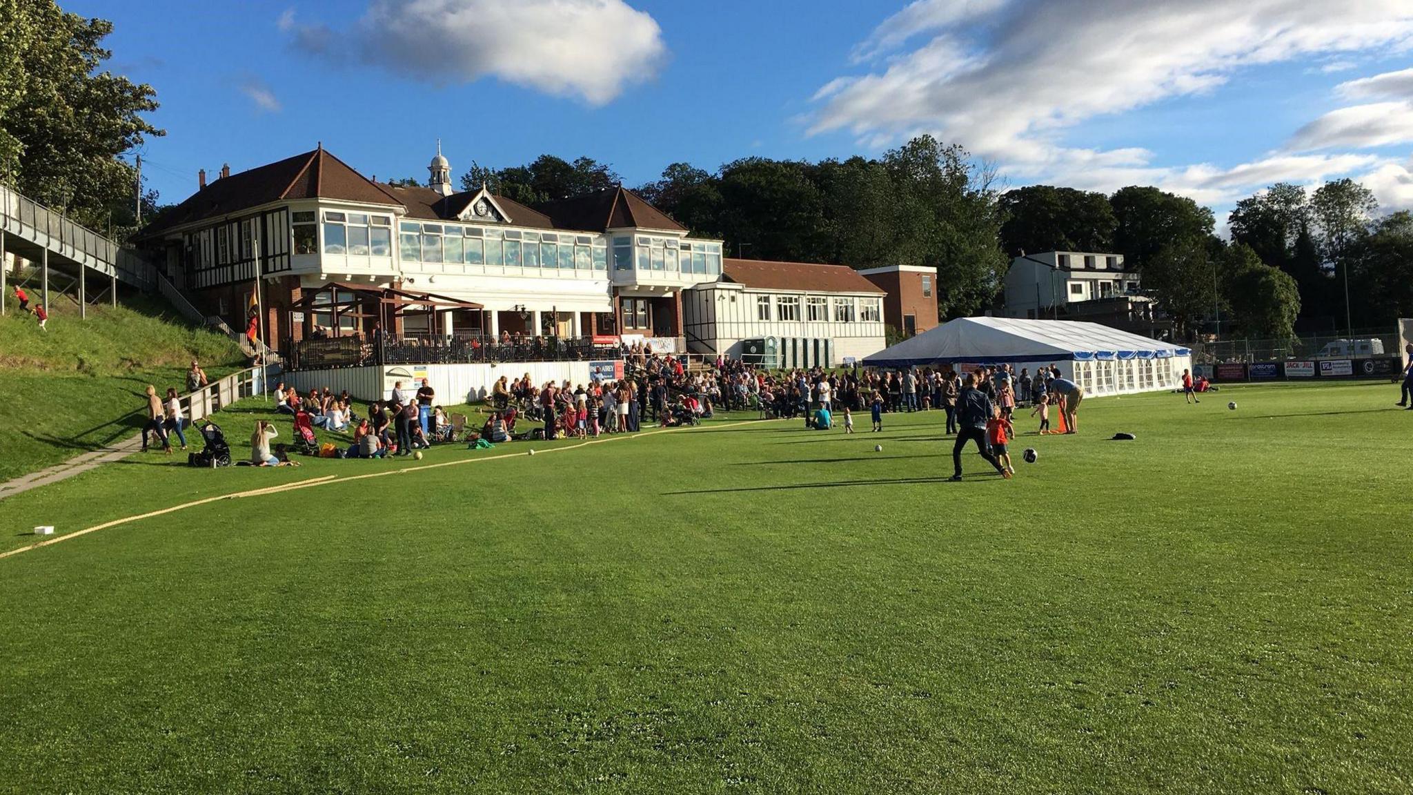 A cricket field with lots of people gathered on the edge with a marquee to the right and a large sports clubhouse with a front balcony  