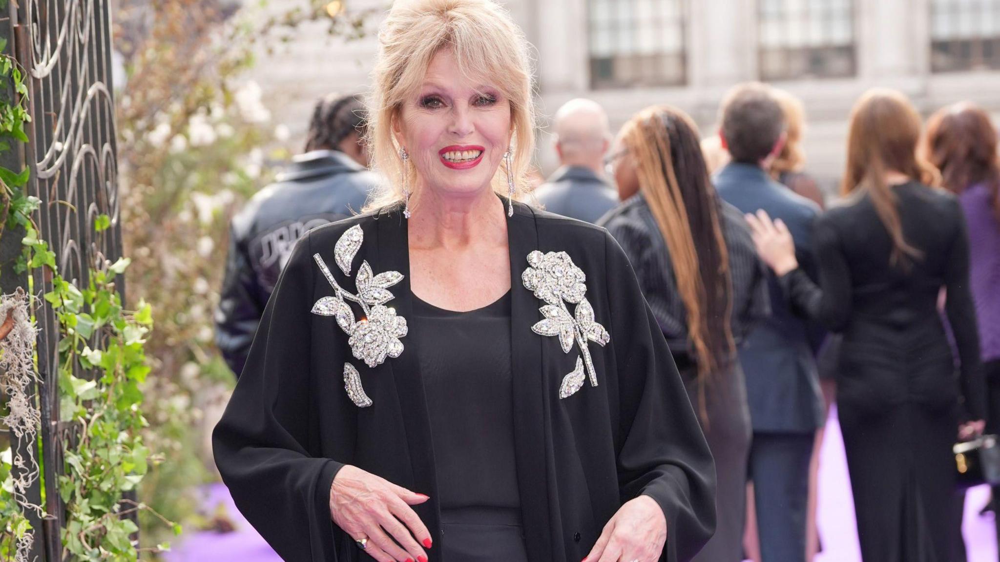 Dame Joanna is walking down a purple carpet outside a building. She is wearing a black embroidered cardigan and has her blonde hair up in a do.