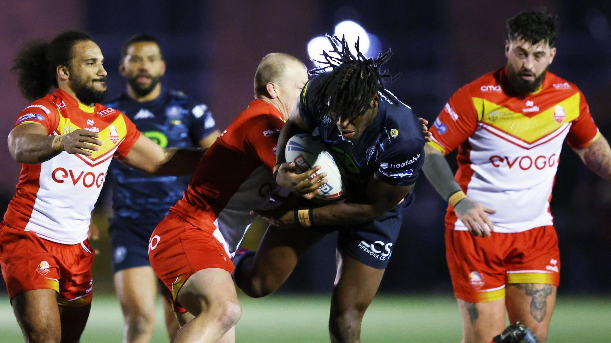 Wigan Warriors forward Junior Nsemba (centre) is tackled by a Sheffield Eagles player with two other Eagles players watching on from either side 