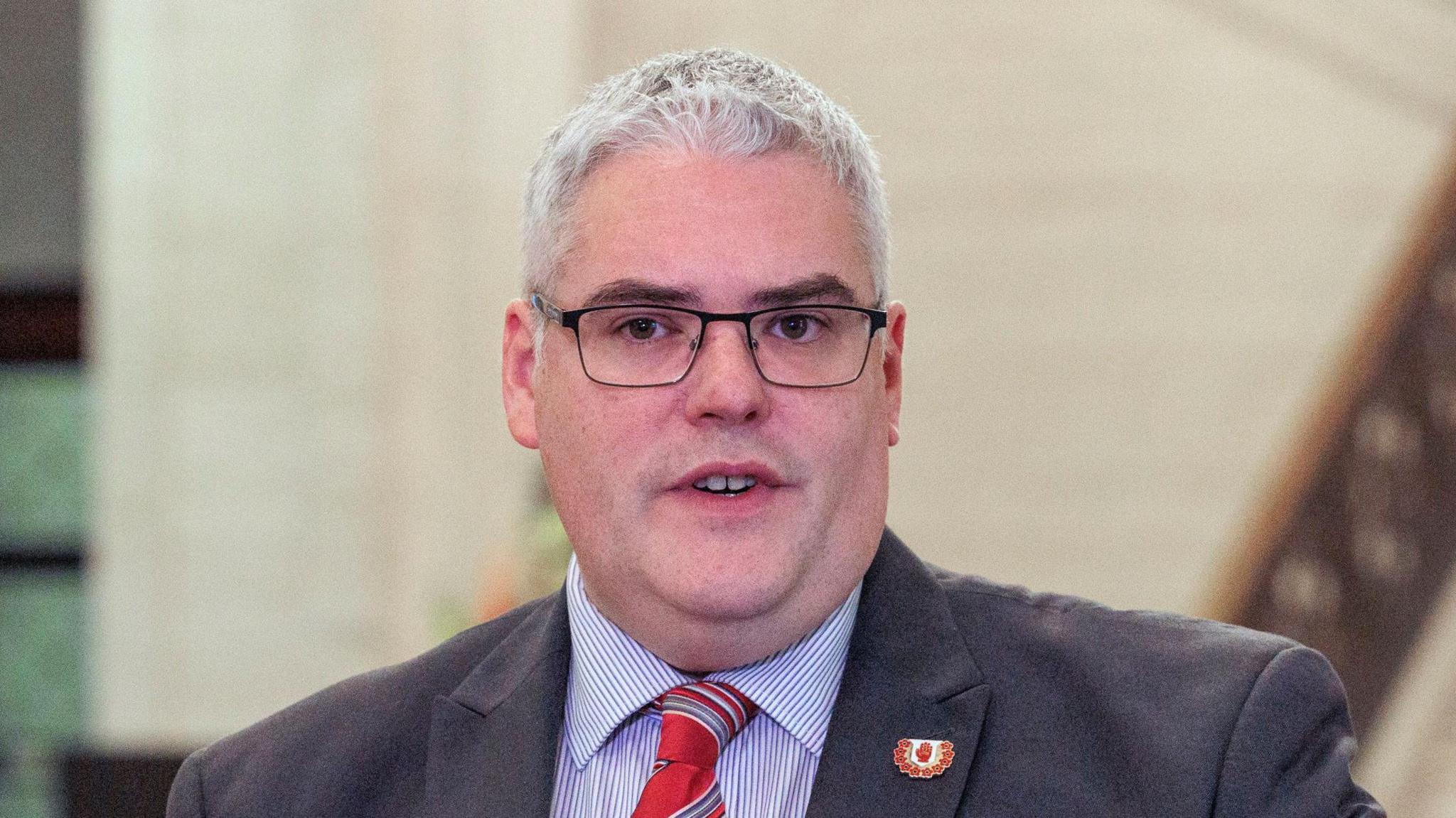 Gavin Robinson has short grey hair and is wearing black rectangular metal glasses, a grey suit, striped shirt and red tie.