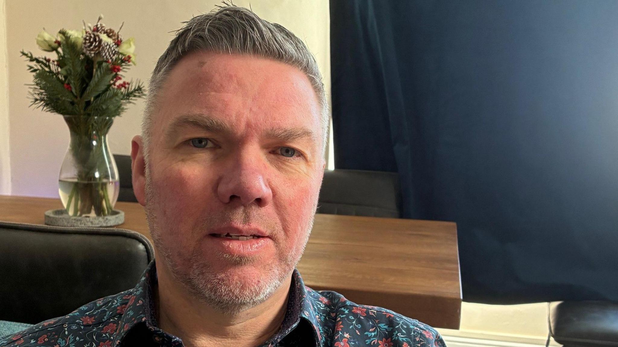 Dave Murphy me një këmishë me lule ulet përpara një tavoline me një vazo me lule sipër.