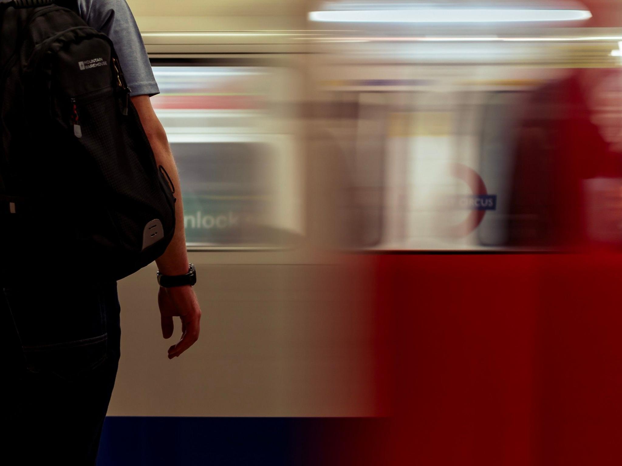 A person wearing a watch on their right wrist stands beside a London underground train which is blurred as it appears to pass by at speed