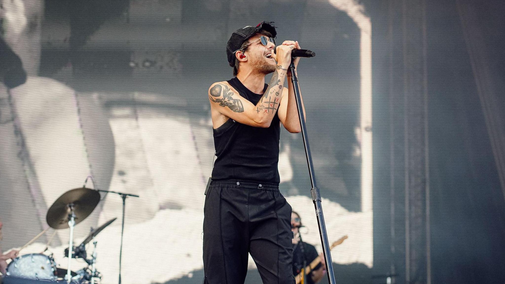 Louis Tomlinson sings on stage, wearing a black cap, black vest, and black trousers