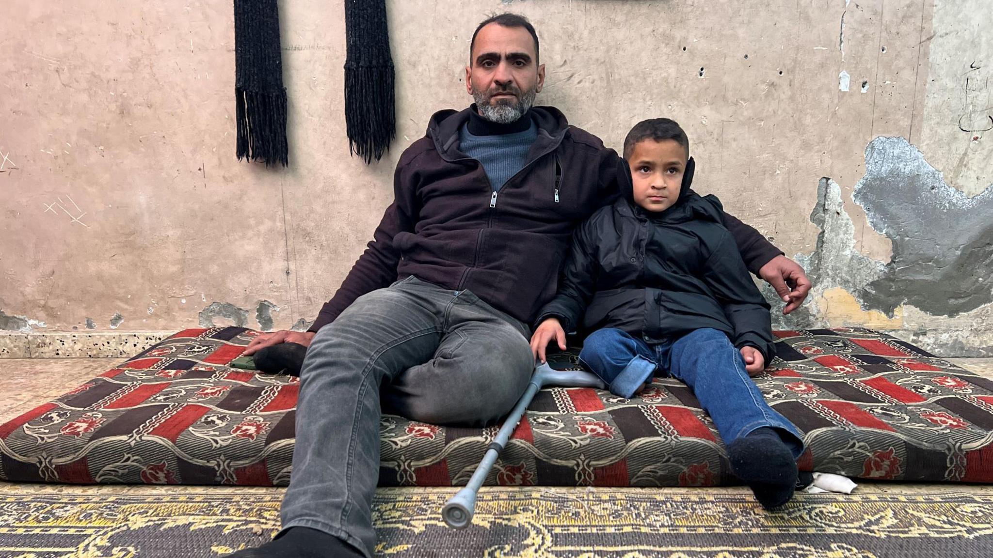 A man and a boy sit on a rug. The boy has a part of his leg missing.