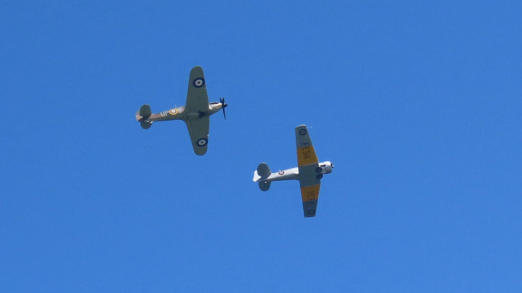 A picture of a Harvard and Hurricane in the sky. There are two planes with the one on the right a grey coloured plane with yellow stripes.
