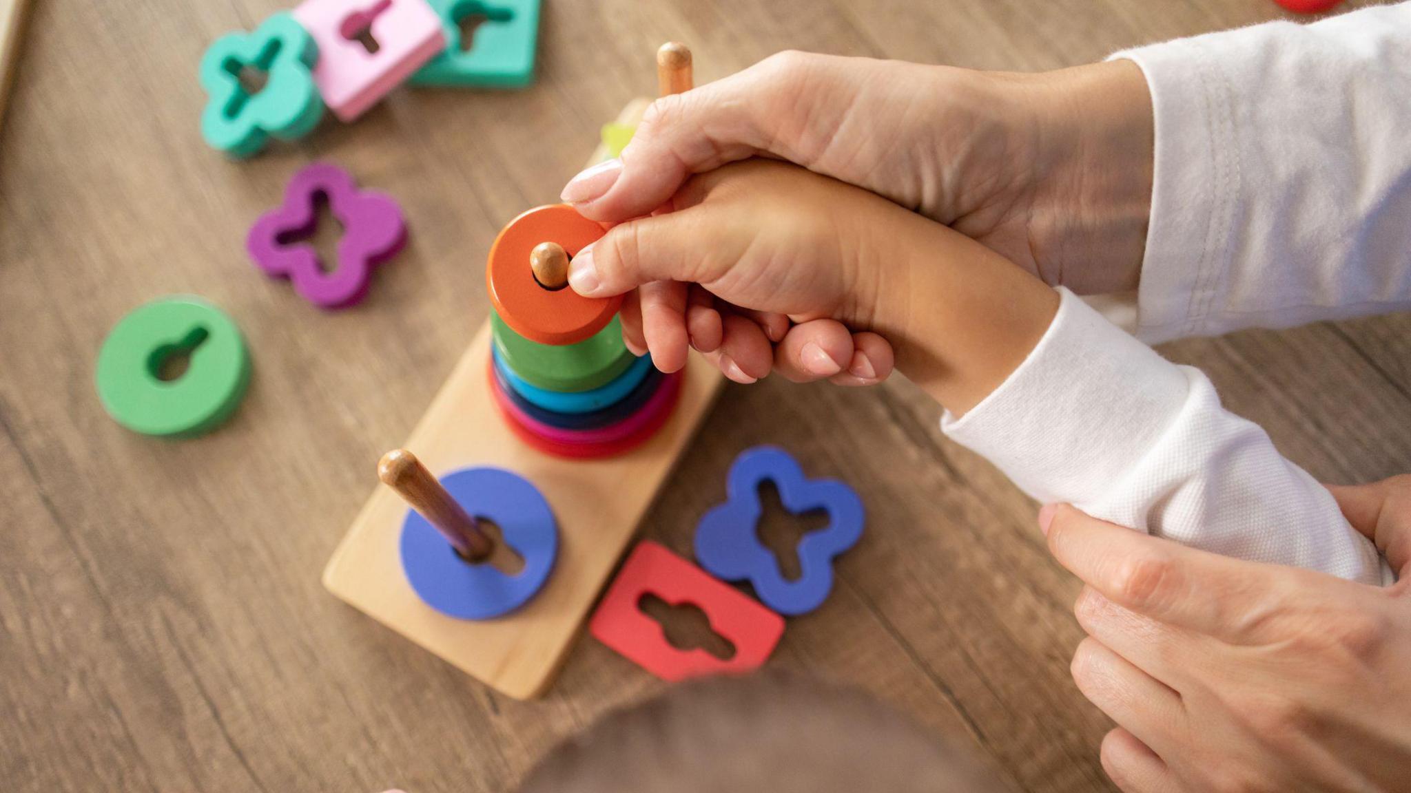 A close-up image of an adult helping a young child place a colourful round piece onto a wooden peg toy. The toy consists of multiple pegs and various brightly coloured, differently shaped pieces scattered around on a wooden surface.