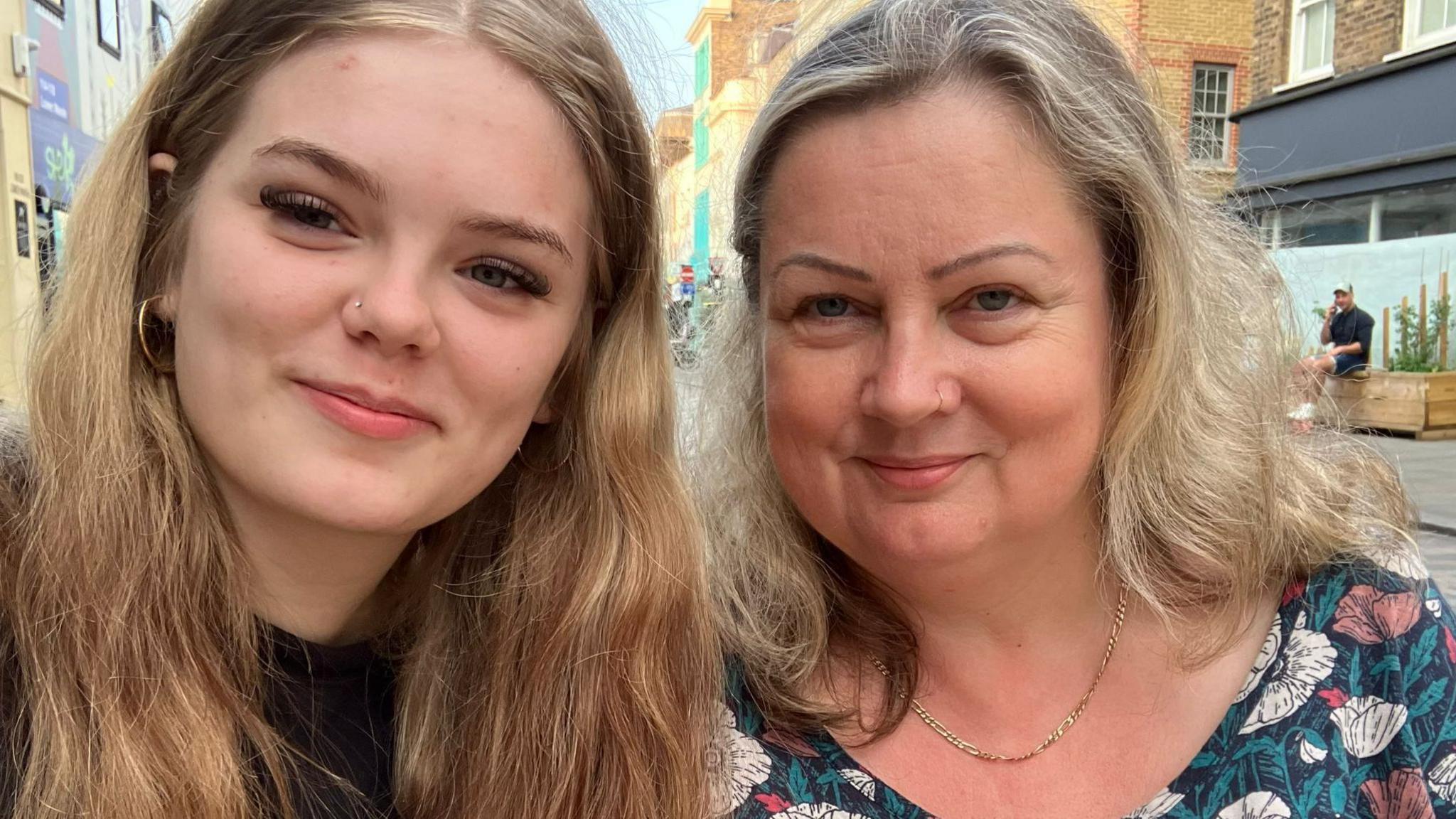 Two women smile at the camera. On the left a teenage girl with blonde hair and on the right a middle aged lady with a floral top smiles at the camera. She also has blonde hair.