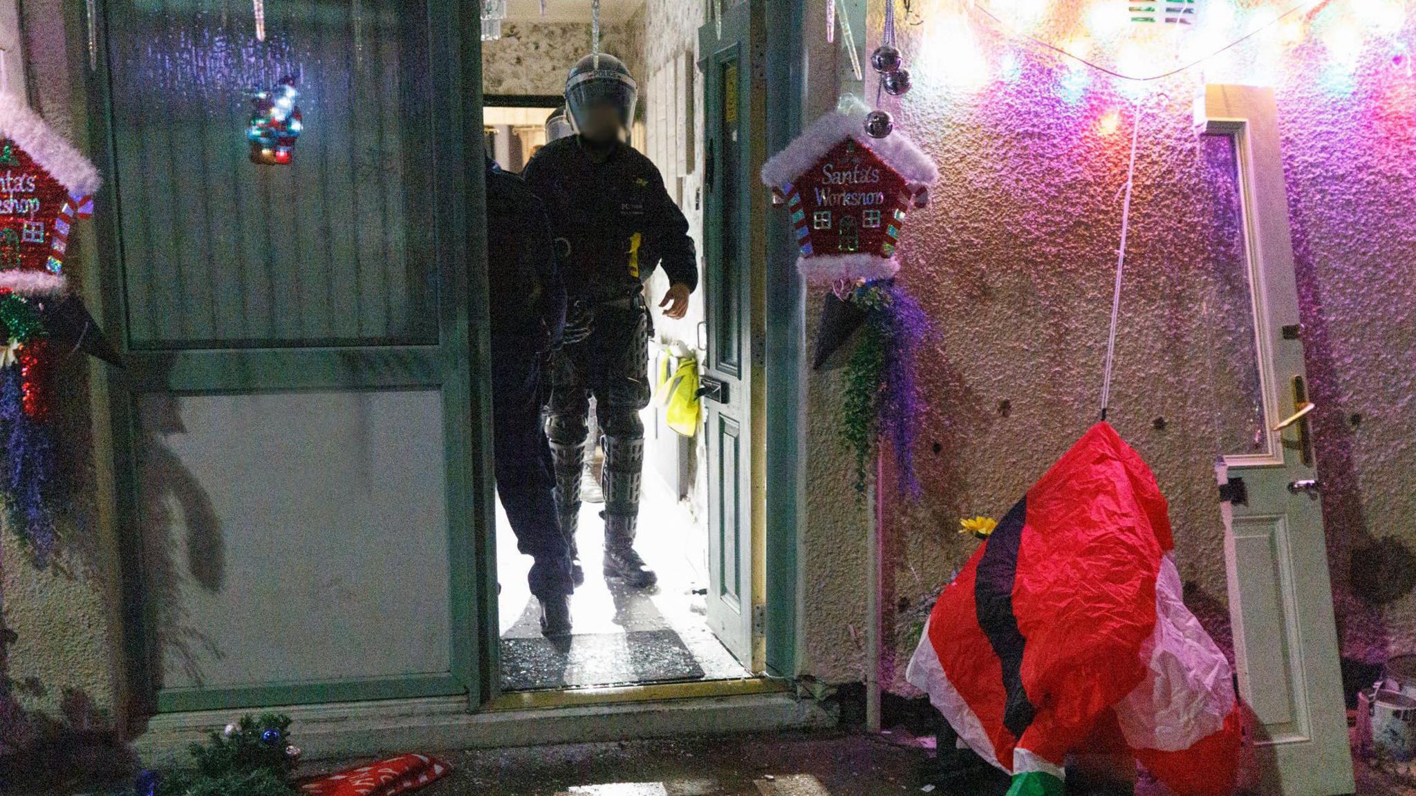 A police officer dressed all in black can be seen walking out a property decked in Christmas decorations. 