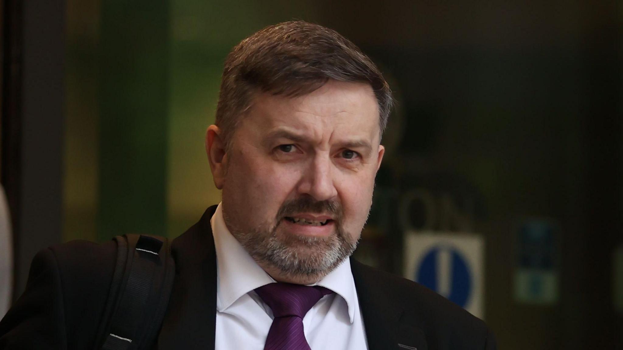 Robin Swann leaving the Clayton Hotel in Belfast after giving evidence to the UK Covid-19 inquiry hearing. He has dark brown hair, wearing a black blazer, white shirt and purple tie carrying a backpack over his shoulder.