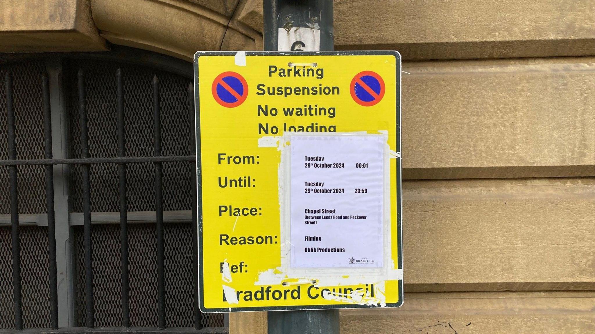 A yellow street sign on a lamppost with a white piece of paper taped over it showing the times when parking will not be available due to filming.  