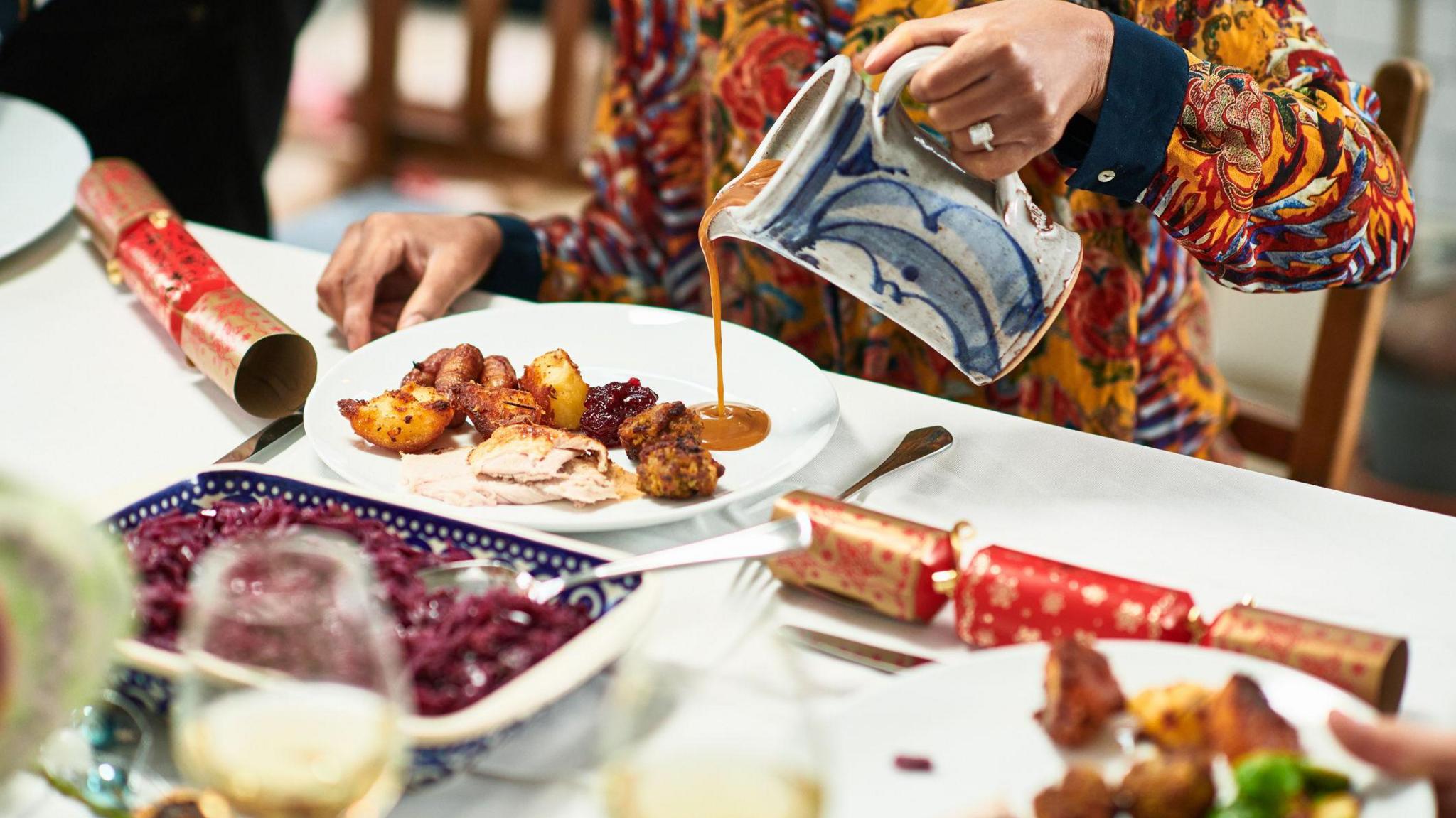 A person wearing a orange jumper with red patterns pour gravy on to a whie plate. The plate has roast potatoes, turkey, and pigs in blankets on it. In front of them is serving dishes and there are two red and gold Christmas crackers on either side of them.