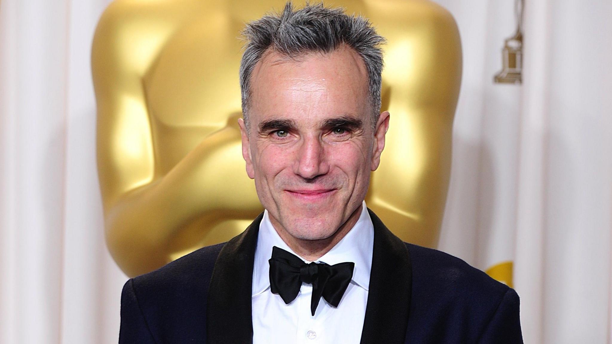 Daniel Day-Lewis, dressed in a dinner suit, holding an Academy Award for Best Actor while standing in front of a large golden Oscar statuette.