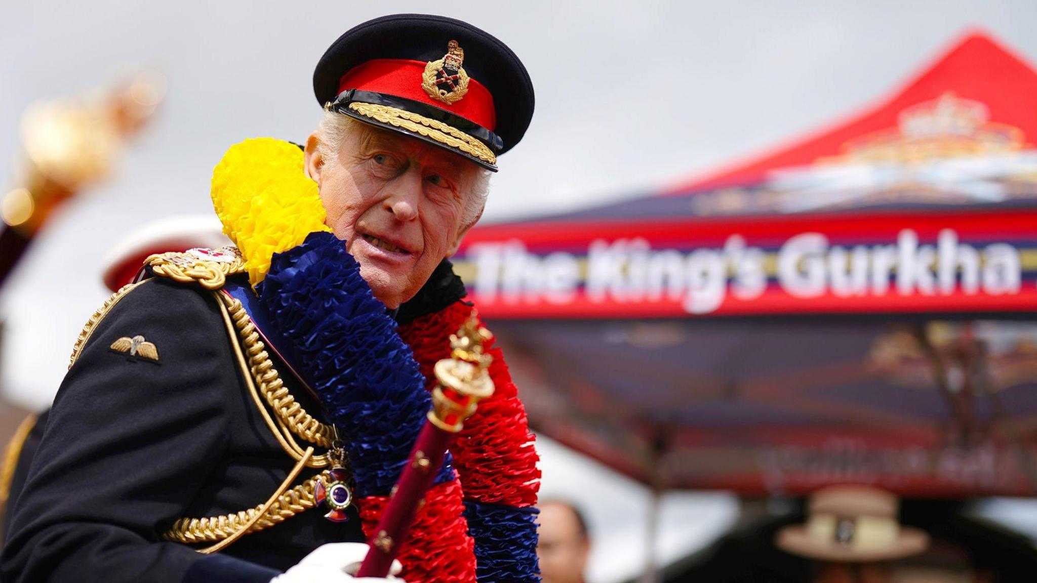 The King in ceremonial military uniform, including hat and gold insignia. He is turning to look at something and has a large garland round his neck with red, yellow and blue.
