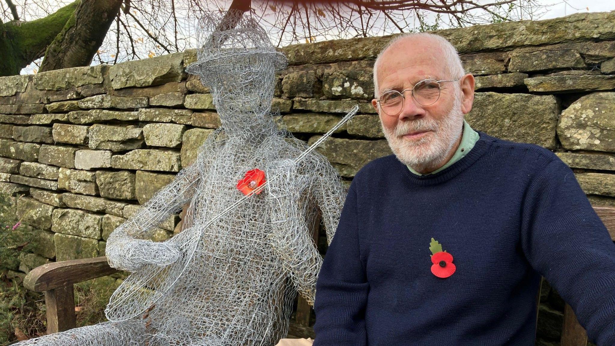 Sam Simson sat on a bench next to a life size human chicken wire model holding a banjo.