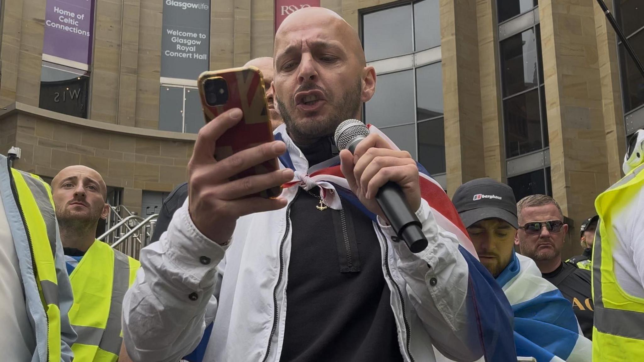 A bald man, Steven Rennie, wearing a white jacket, with a Union flag draped round his shoulders, delivers a speech to a crowd. He is flanked by male supporters, some of whom are wearing fluorescent yellow vests. He is holding a microphone in his left hand and reading from a phone, which has a Union Jack protective cover on it. The steps to the Glasgow Royal Concert Hall can be seen in the background.  