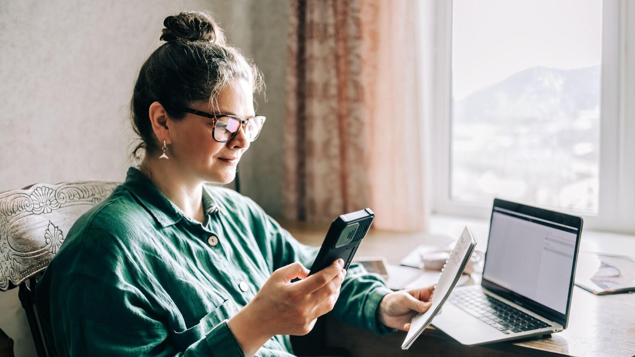 Woman sits at a window seat holding a phone and a pad with a computer on a table in front of her.
