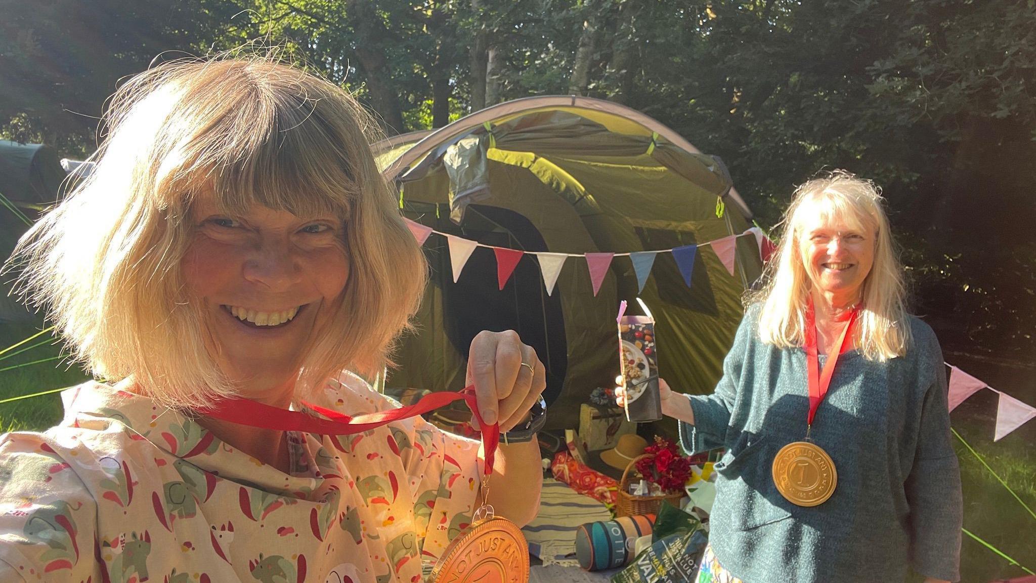 Carey Fluker is in the forefront of the photo wearing a patterned shirt and with a big medal around her neck. She has a blonde bob and is about 60. Behind her you can see another woman and a tent.