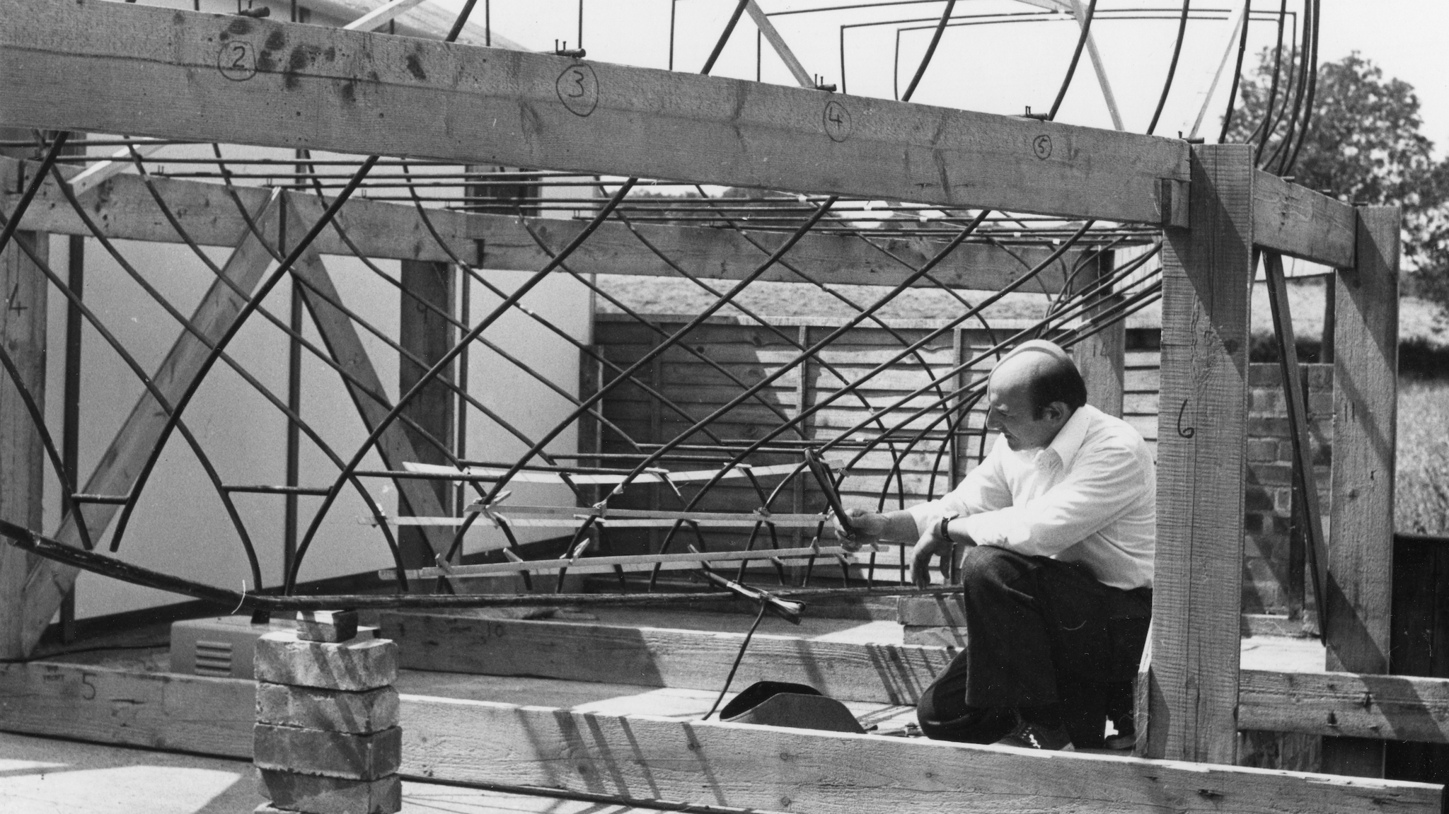 A black and white image of Lee Holmes wearing a shirt and trousers and looking at a partial frame of a boat