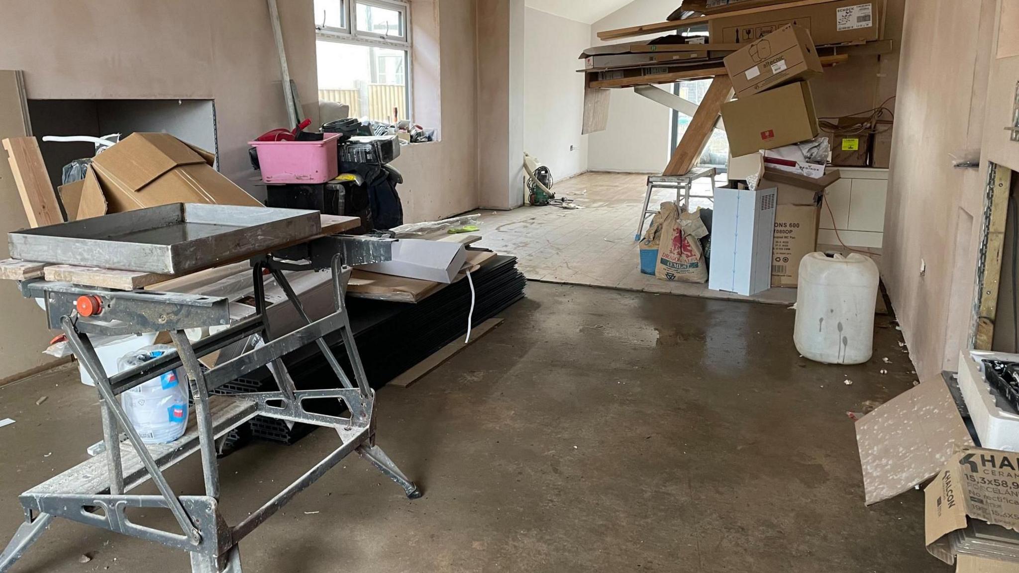 A front room with no carpet. A decorating table stands in the middle of the room and boxes are piled up in one corner.