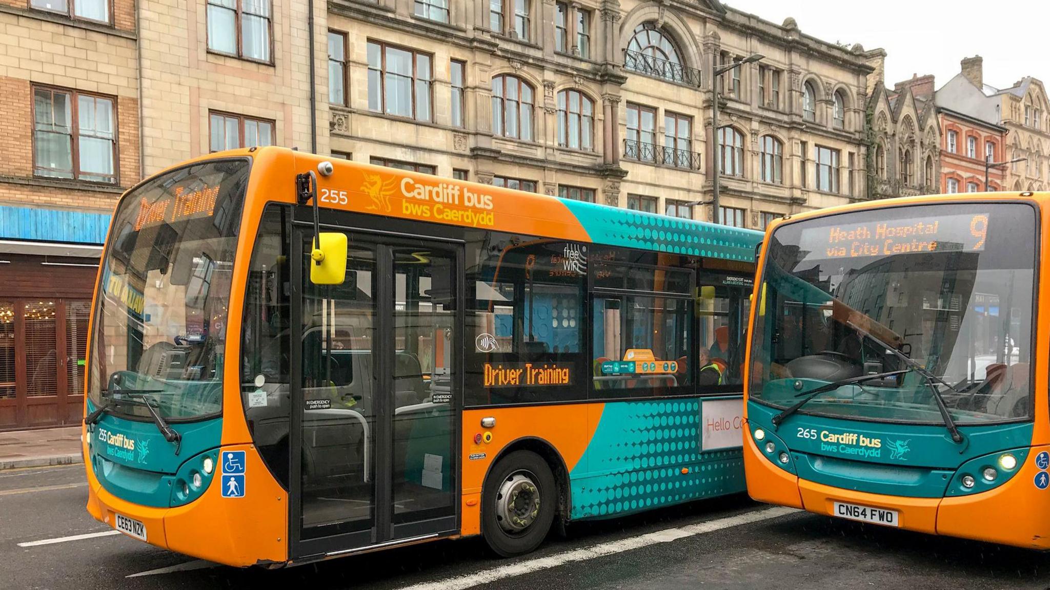 Cardiff bus being used for driver train passing another one of the company's buses in the city centre. Tall buildings can be seen in the background.