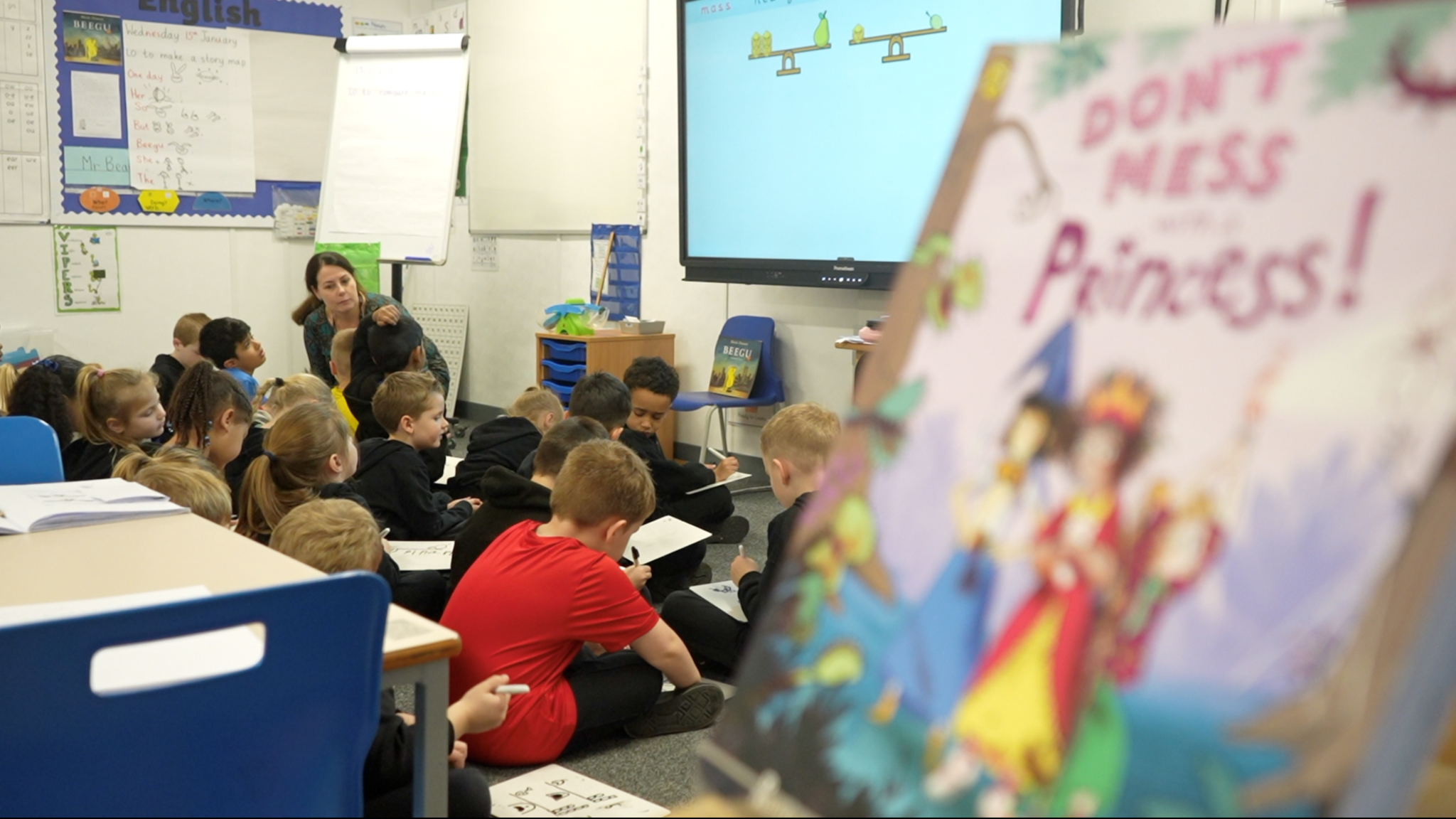 Children are sat on the floor of a classroom and drawing on whiteboards. Their teacher is kneeling while instructing them.