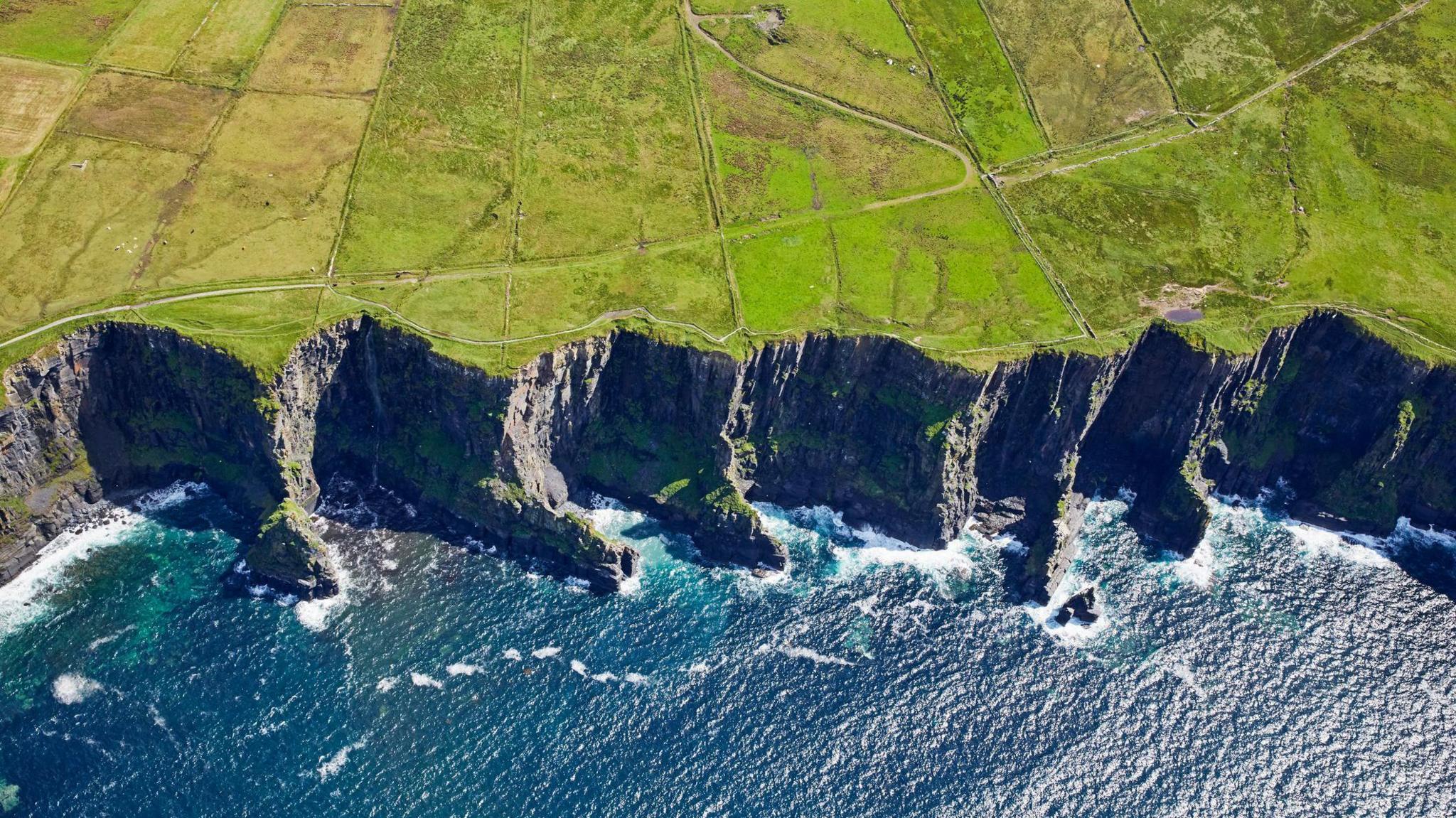 An aerial view of the Cliffs of Moher in County Clare on the west coast of Ireland. Waves are gently breaking below the tall cliffs which stretch over a large distance. The ground behind the cliffs is fields crossed by several paths and trails.