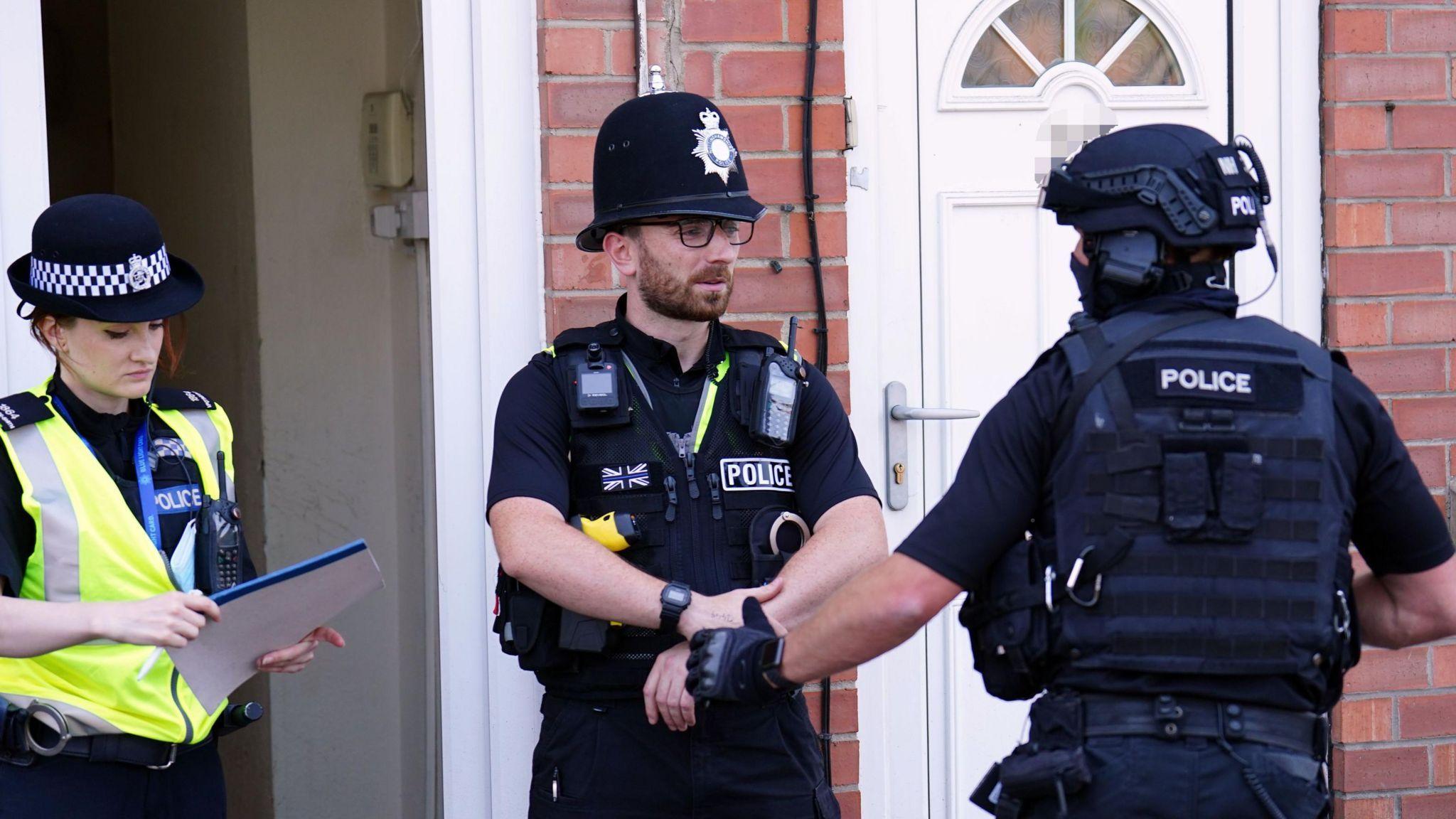 Three police officers wearing their uniforms and standing outside a red brick house. One female officer on the left is holding a document, a male officer in the middle is standing with his hands together and speaking to another male officer on the right, who has his back to the camera