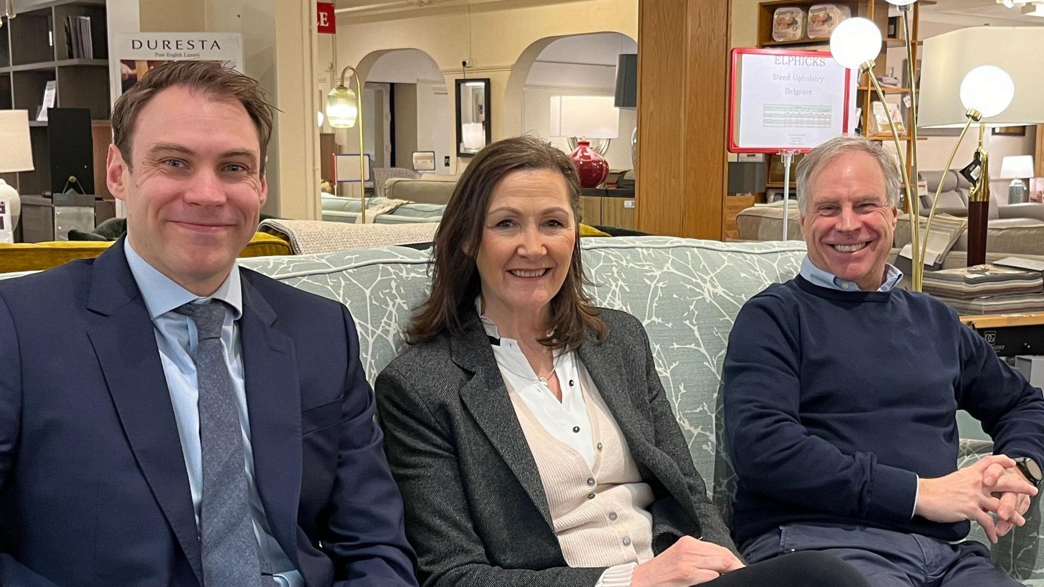 Jonathan, Sue and Kim are all sat on a blue couch inside the store. They are smiling and looking at the camera. Jonathan wears a navy blue blazer, light blue shirt and faded blue tie. Next to him is Sue. She wears a grey blazer, cream cardigan and white blouse. She has brown shoulder length hair and wears a silver necklace. Kim is wearing a navy blue jumper, light blue shirt and has his hands clasped in front of him. He has a watch on his left wrist. 