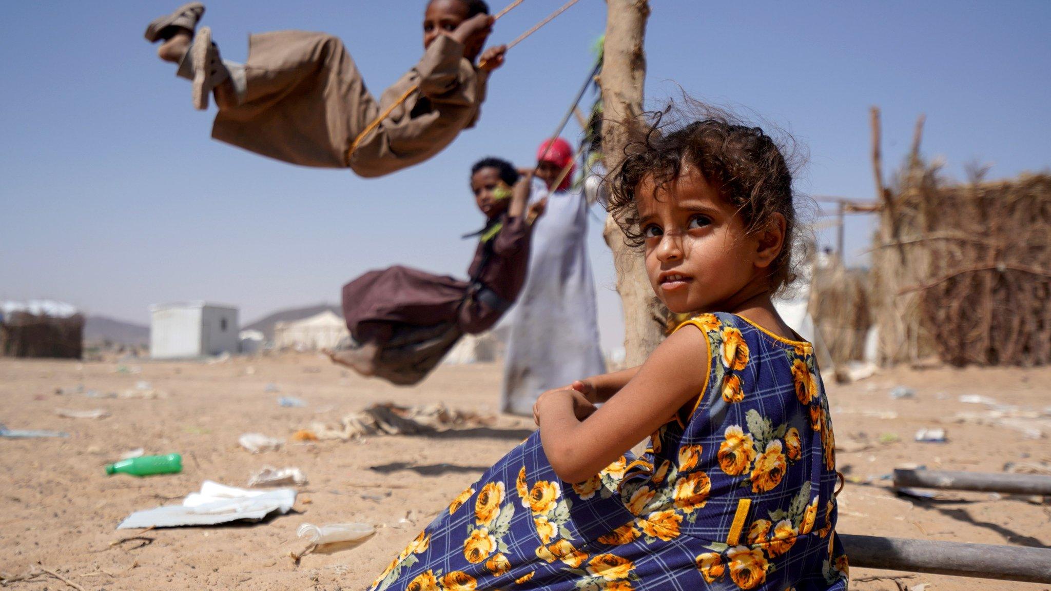 A girl sits as other children play on swings at a camp for internally displaced people in Marib, Yemen (16 February 2021)